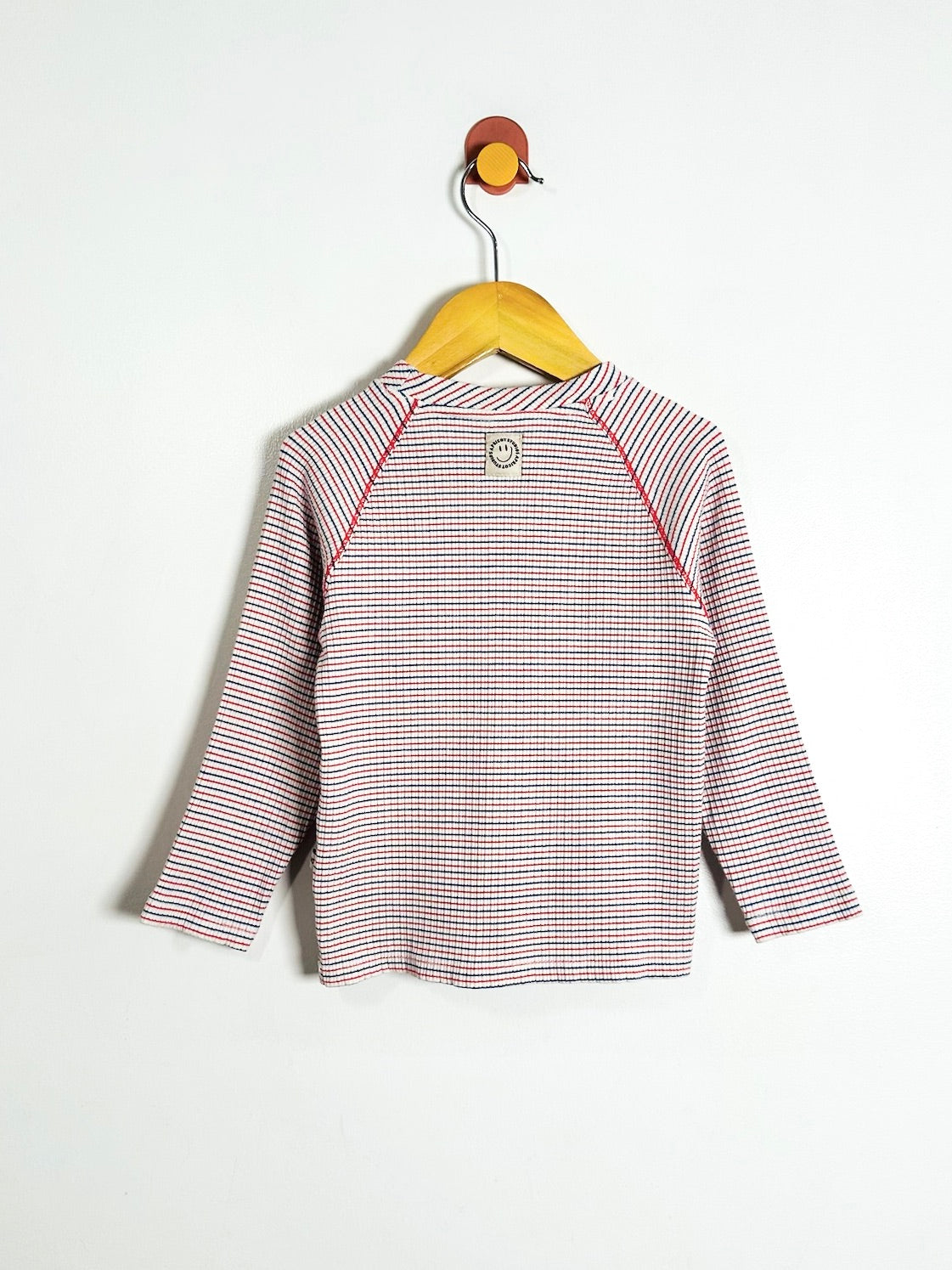 A children's long-sleeve top with thin red, blue, and white horizontal stripes, hanging on a wooden hanger against a plain white wall.