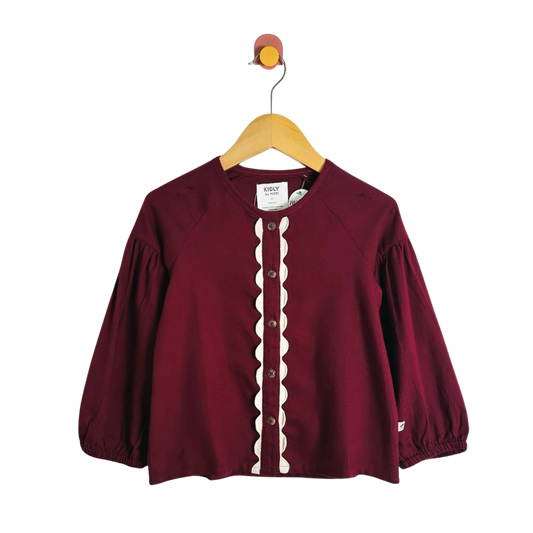 A burgundy long-sleeve raglan top with a cream scalloped placket and button front, hanging on a wooden hanger against a white background.