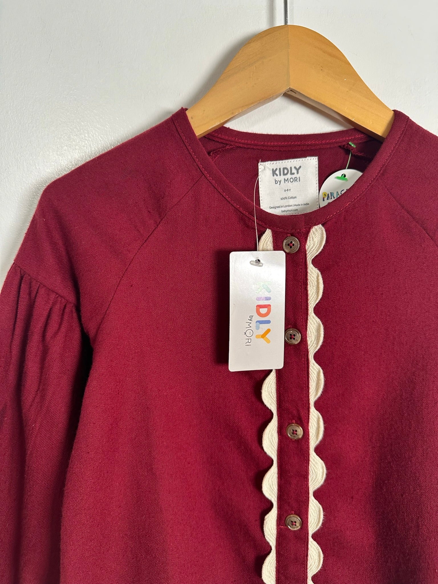 A kids' long-sleeve top in deep burgundy, featuring a cream scalloped trim down the front button placket. The top is displayed on a wooden hanger with a white 'KIDLY by MORI' brand tag and a smaller 'DIY' hangtag attached.