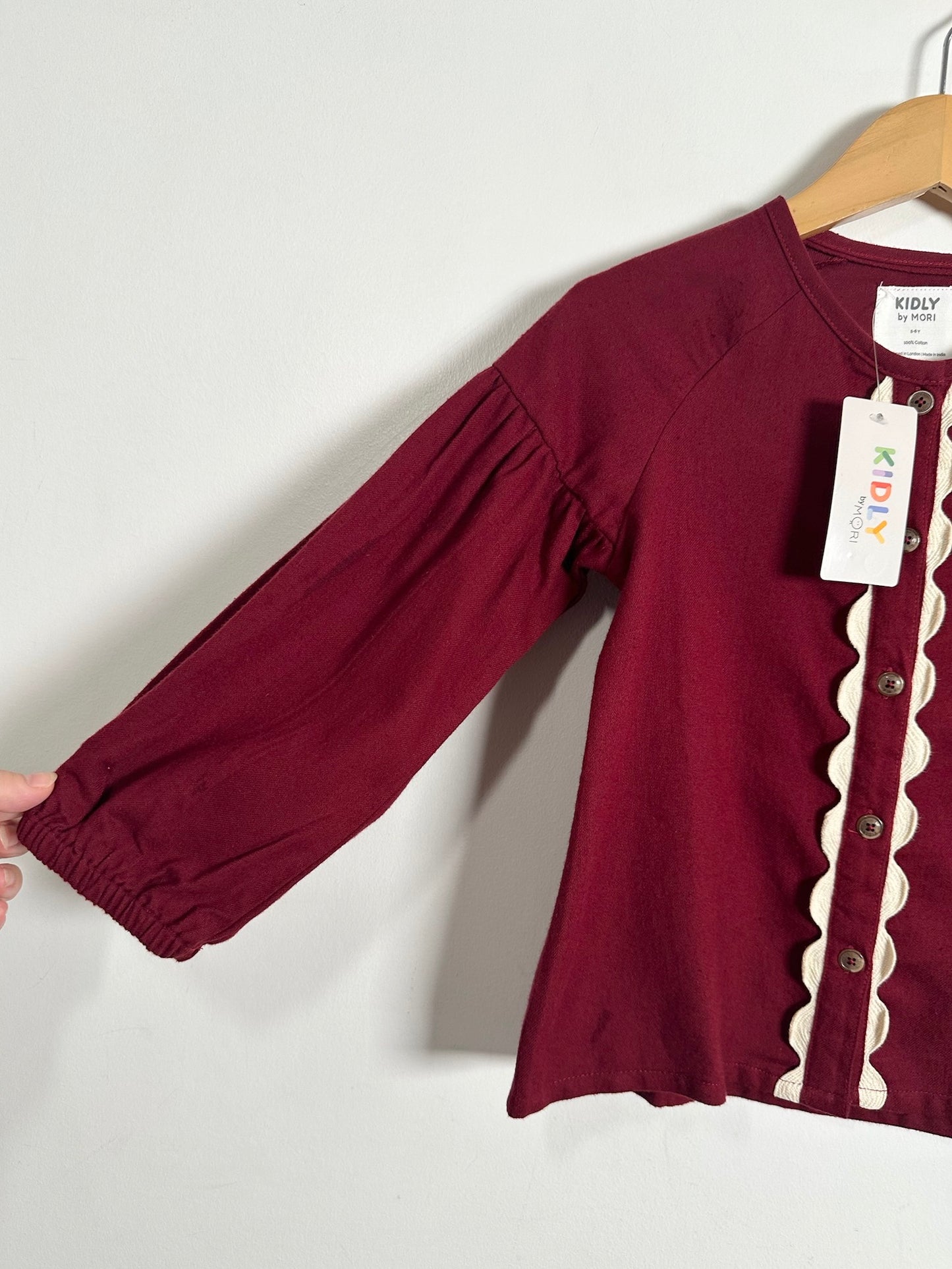 A kids' long-sleeve top in deep burgundy, hanging on a wooden hanger against a plain light gray wall. The shirt features a scalloped cream trim down the front button placket and a white brand label that reads 'KIDLY KIDS'.