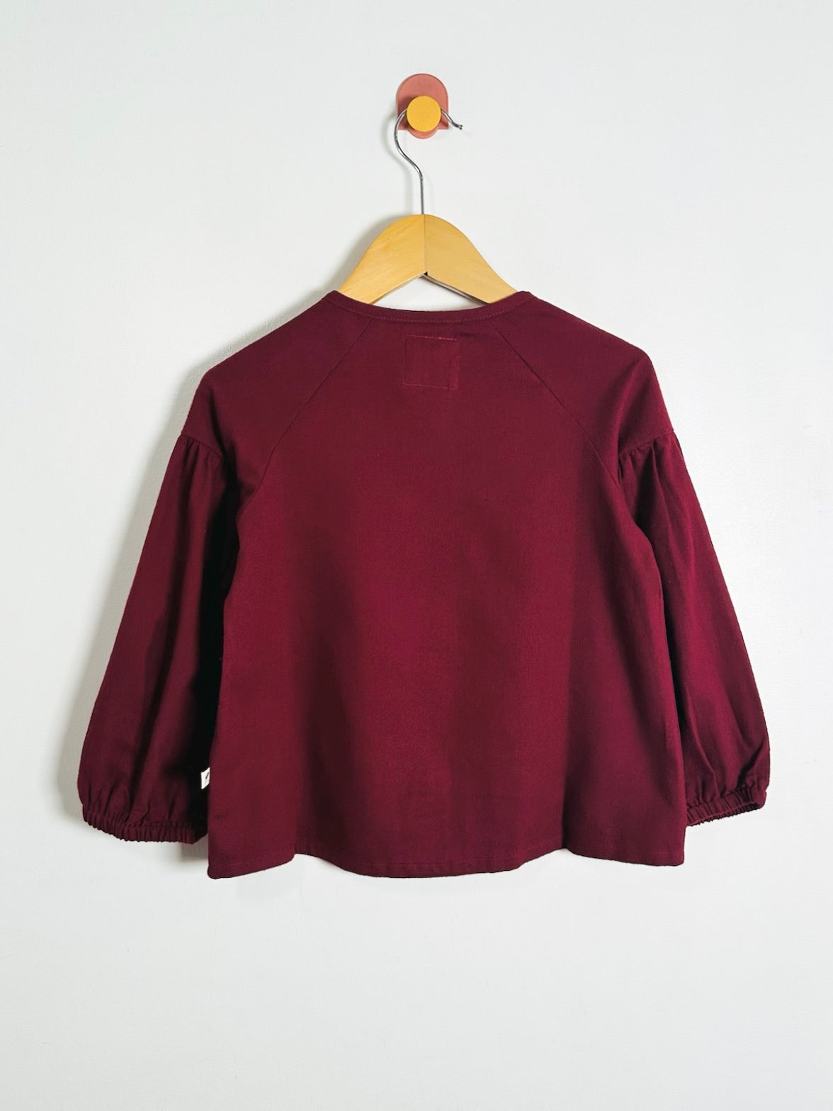 A burgundy long-sleeve crop top with gathered elbow-length sleeves, hanging on a wooden hanger against a plain white wall.