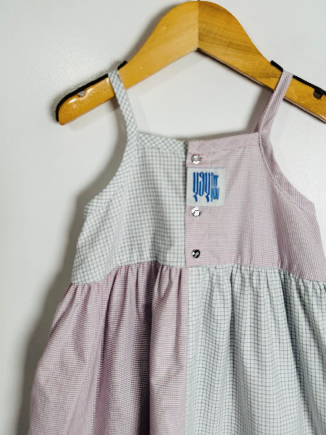 A sleeveless baby dress with a split gingham pattern, featuring a light pink and white checkered lower half and a white and light gray checkered upper half. It is hanging on a wooden hanger against a plain white wall.