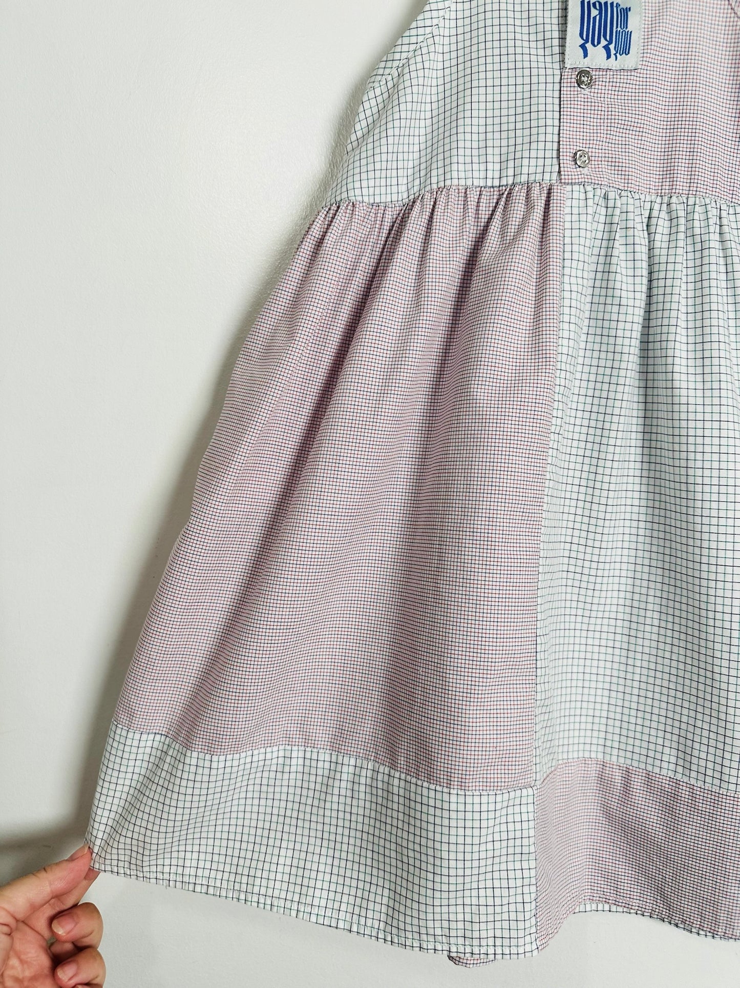 A close-up of a patchwork dress featuring a dusty pink gingham skirt, a white and light blue gingham bodice, and a hand holding the hem to show its flexibility.