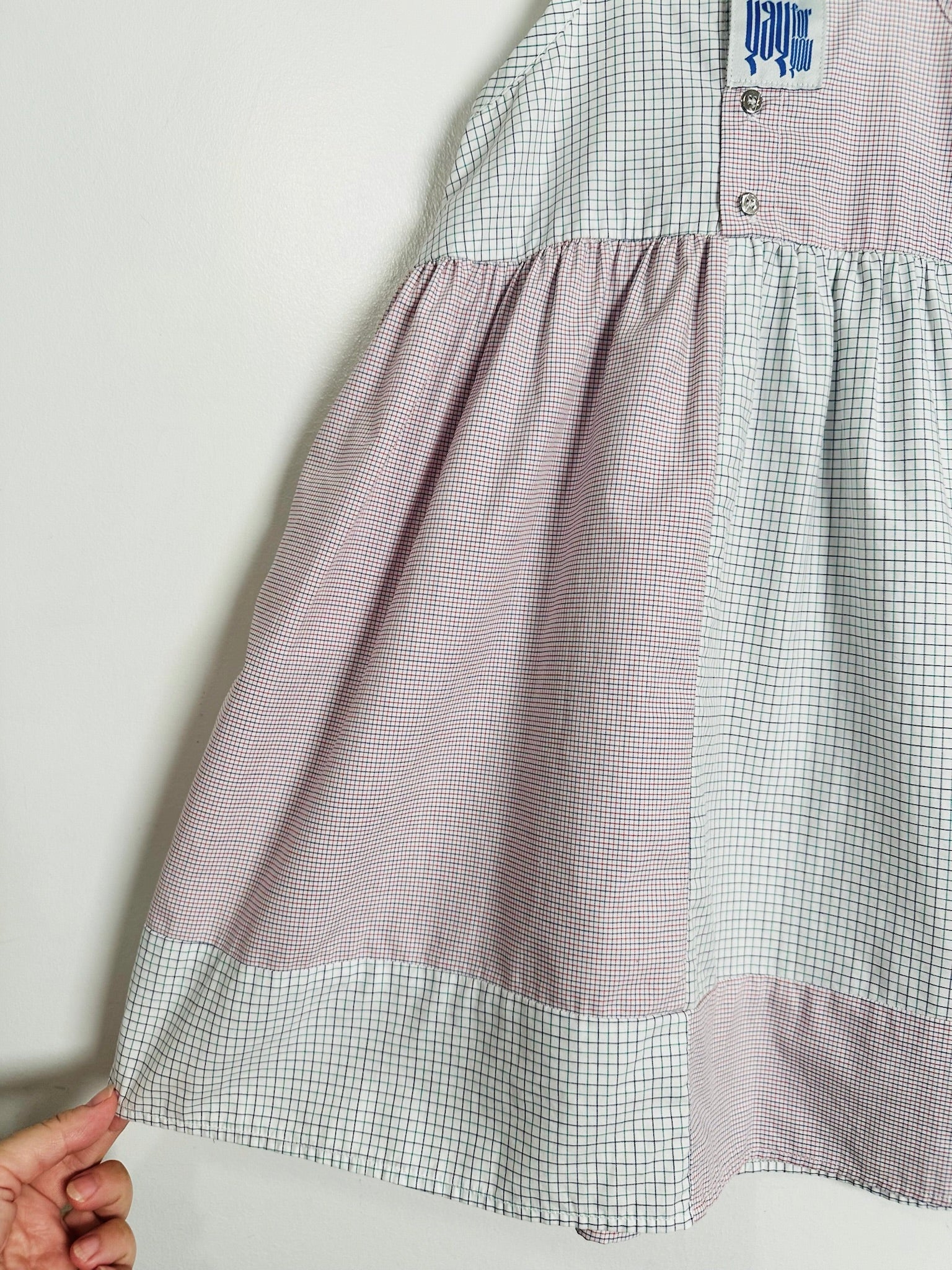 A close-up of a patchwork dress featuring a dusty pink gingham skirt, a white and light blue gingham bodice, and a hand holding the hem to show its flexibility.