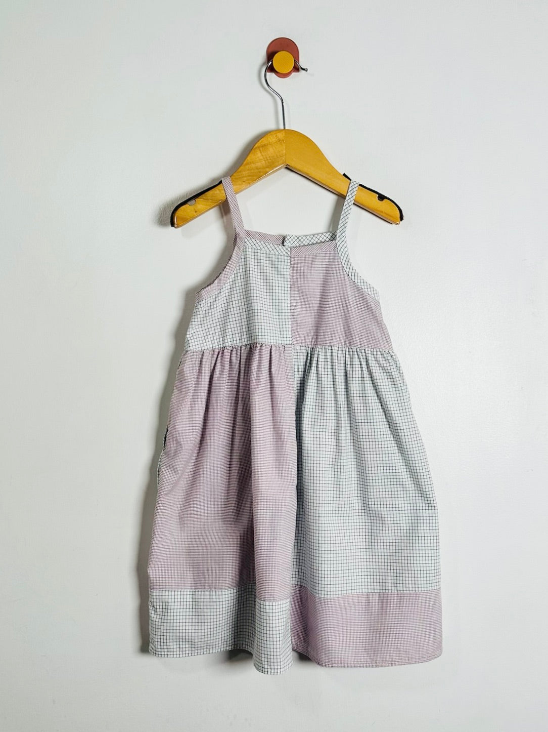 A sleeveless baby dress with a patchwork design of dusty lavender and white gingham, hanging on a wooden hanger against a plain white wall.