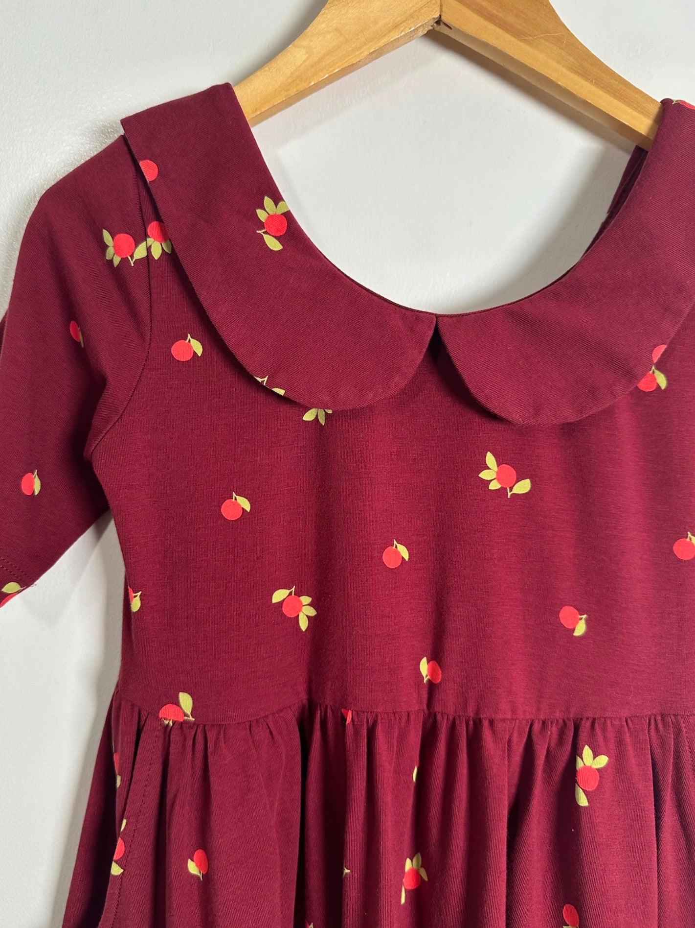 A close-up of a dark red dress with a small red and yellow strawberry print, featuring a Peter Pan collar and short sleeves, hanging on a wooden hanger against a white wall.