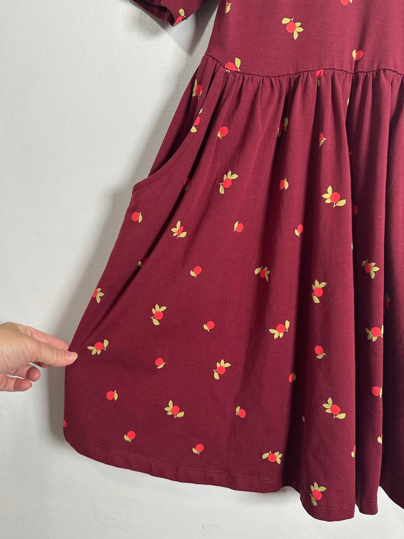 A close-up of a burgundy dress with a small red and yellow strawberry print, featuring a side pocket and a hand pointing to the fabric.
