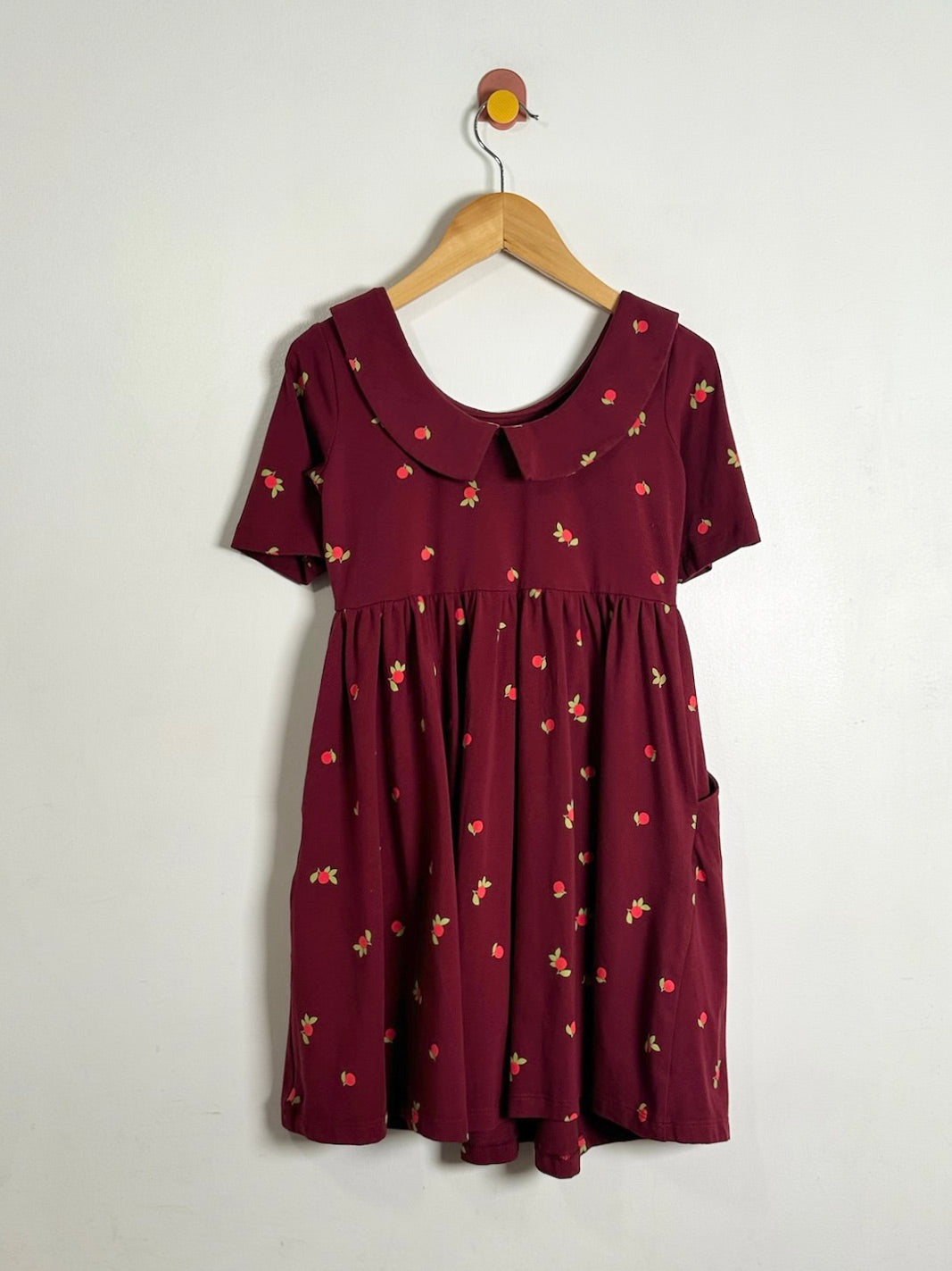 A short-sleeve babydoll dress in deep burgundy with a small red and yellow strawberry print, hanging on a wooden hanger against a plain off-white wall.