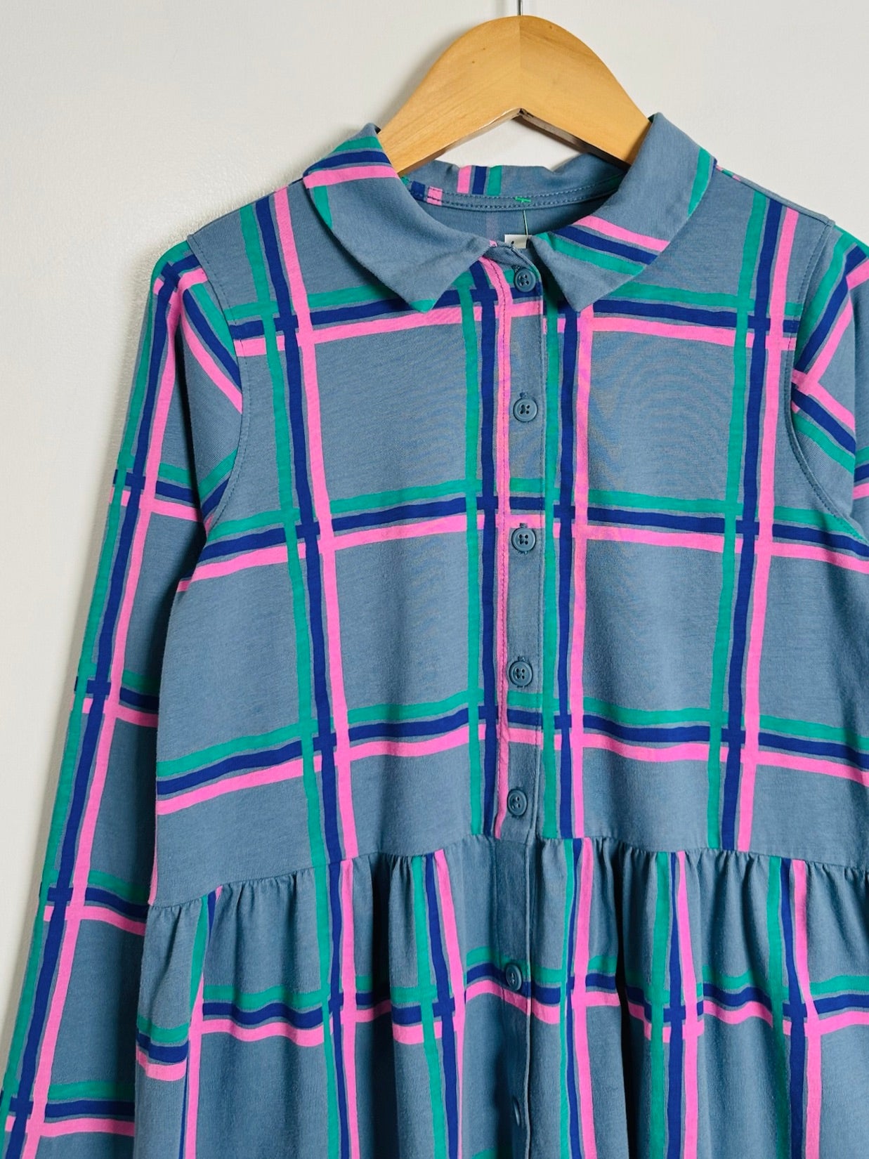 A long-sleeve collared button-front dress in a blue-gray base with a plaid pattern of bright pink, teal green, and dark navy lines, hanging on a wooden hanger against a plain light background.