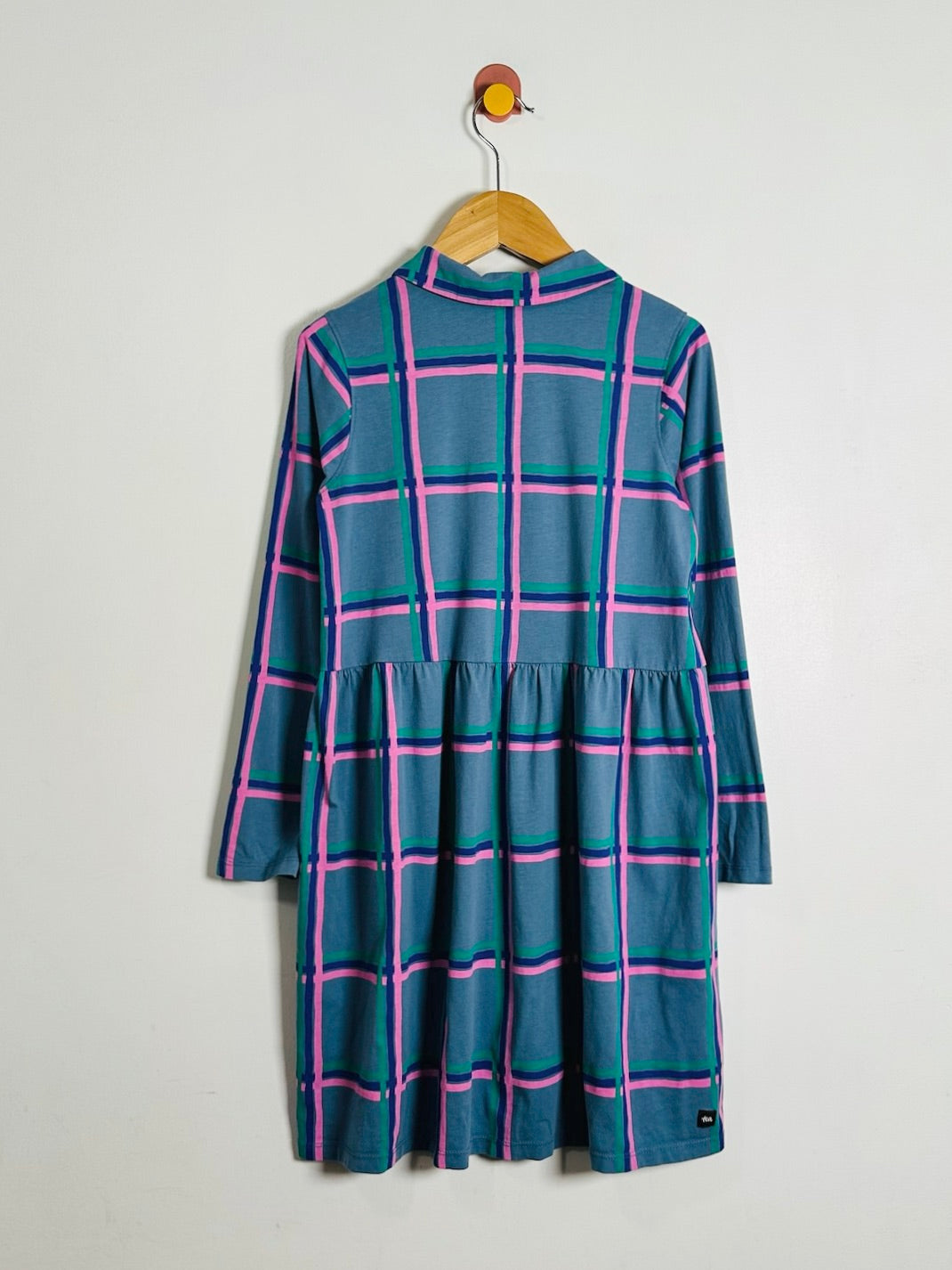 A long-sleeve high-neck dress with a teal blue base and a pink and green plaid pattern, hanging on a wooden hanger against a plain light gray wall.