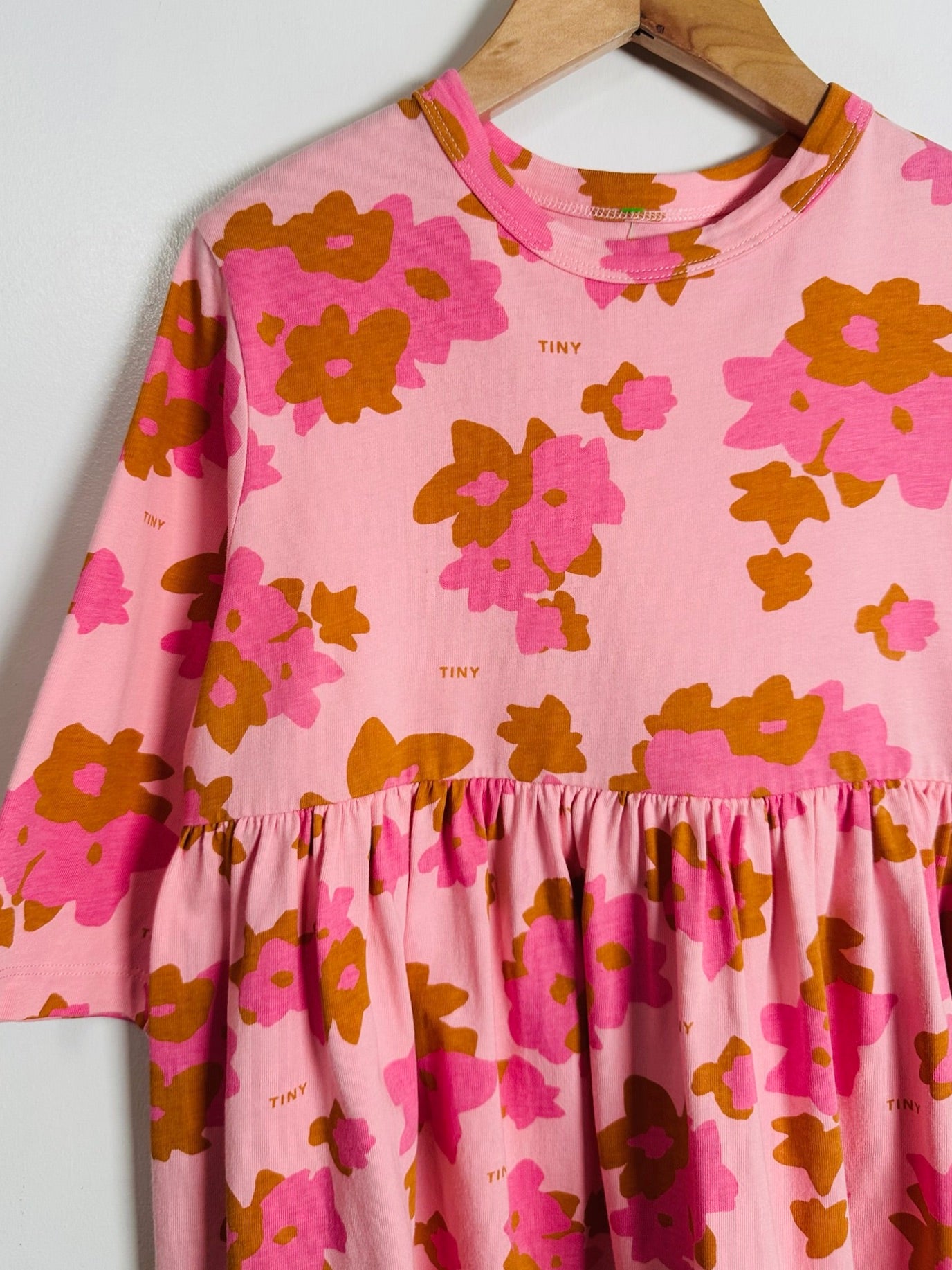 Pink tiered dress with a retro floral pattern in hot pink and orange-brown, hanging on a wooden hanger against a light gray wall.