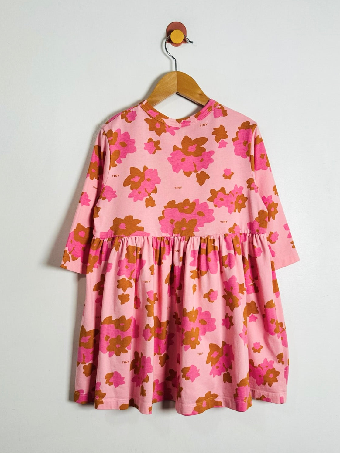 A pink long-sleeve dress with a brown and magenta floral pattern, hanging on a wooden hanger against a plain light gray wall.