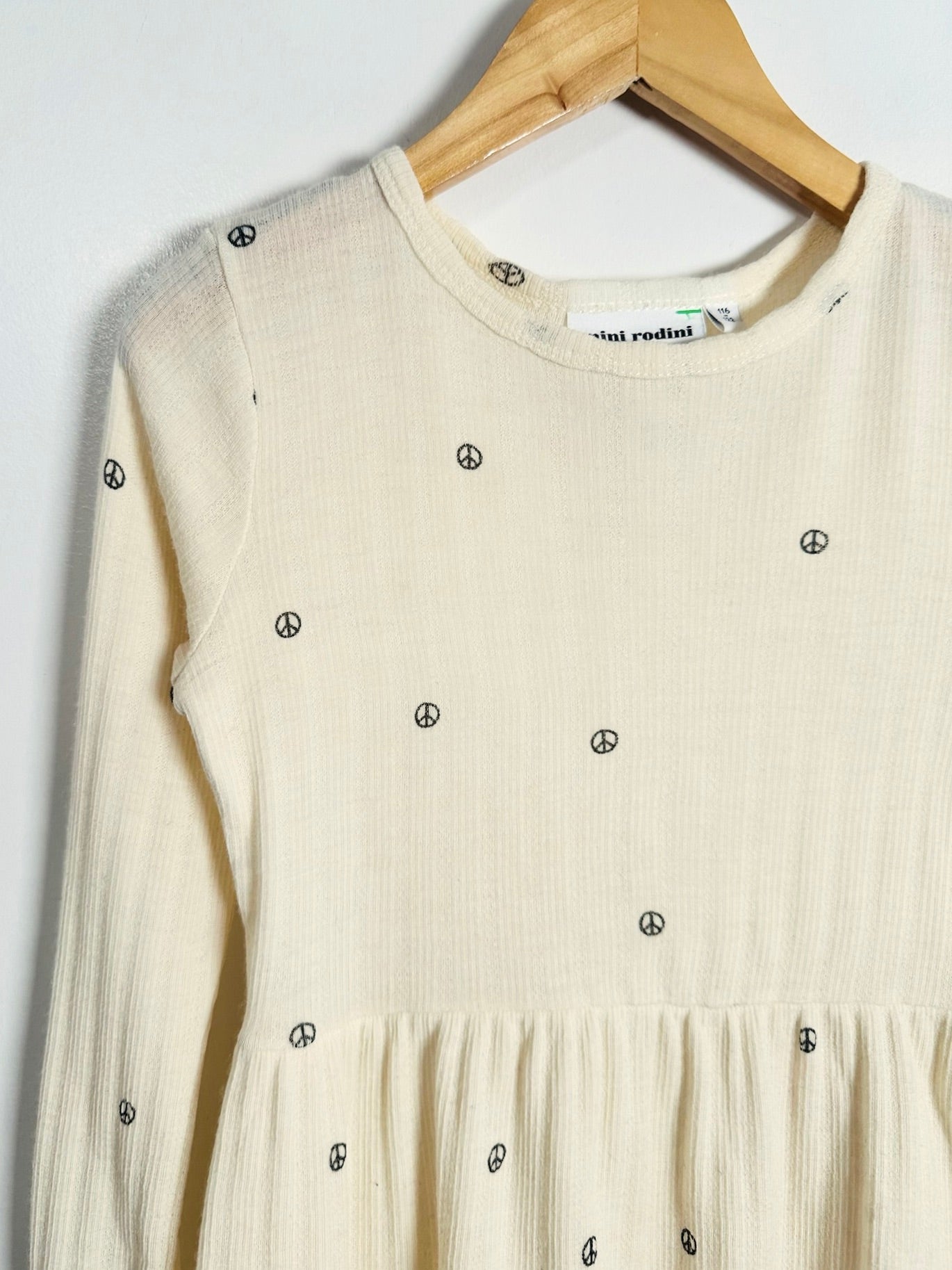 A cream-colored long-sleeve ribbed knit dress with small black peace sign embroidery, hanging on a wooden hanger against a plain white wall.