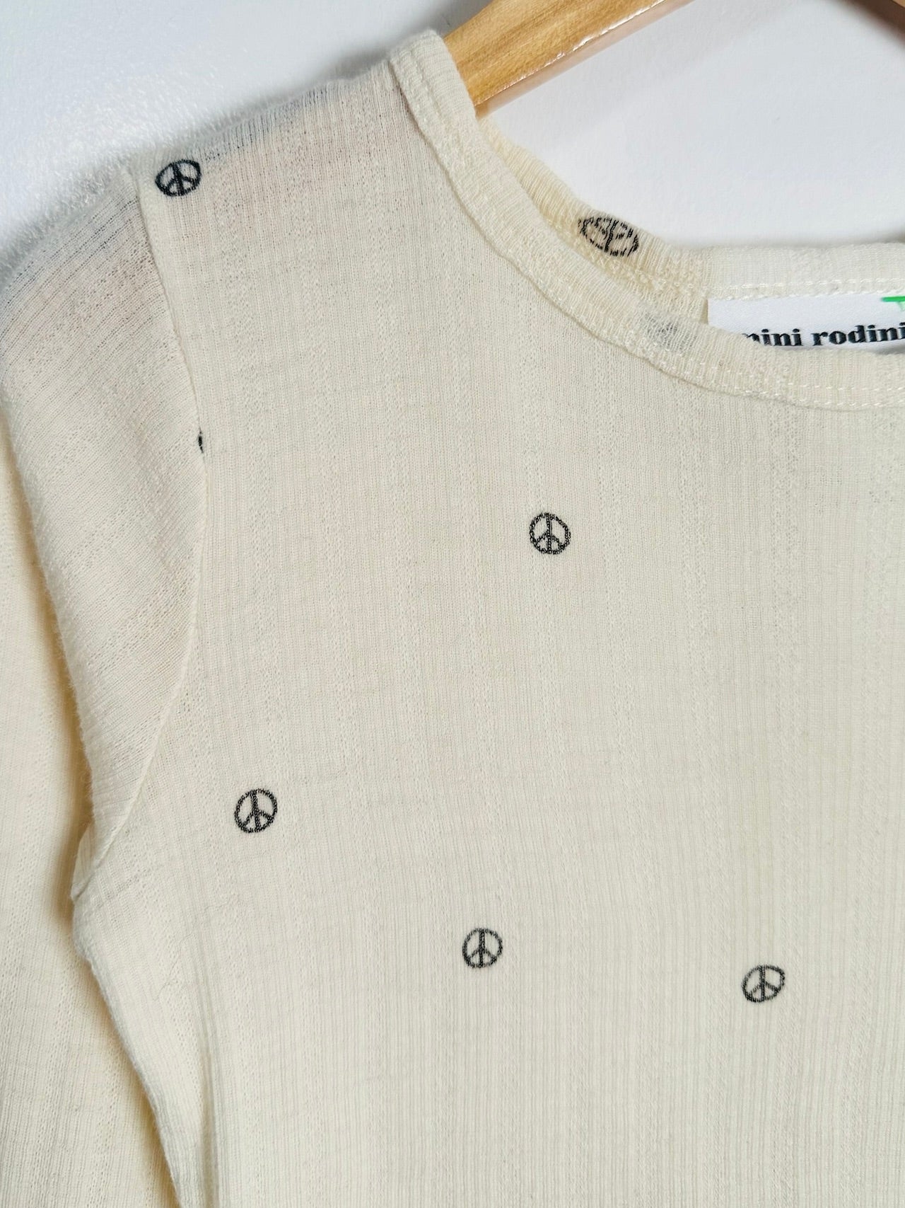 A close-up of a cream-colored ribbed knit sweater with small black peace sign motifs, hanging on a wooden hanger against a white background.