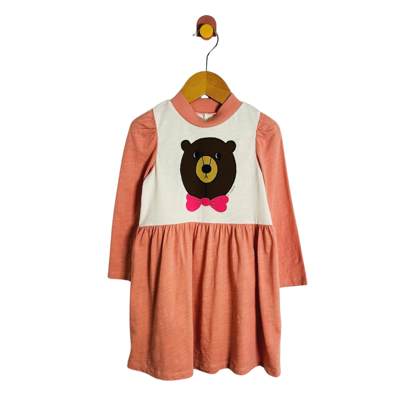 A toddler girl's long-sleeve dress with a terracotta orange skirt and sleeves, and a cream-colored bib-style chest panel featuring a cartoon bear face with a pink bow tie. The dress is hanging on a wooden hanger against a plain white background.
