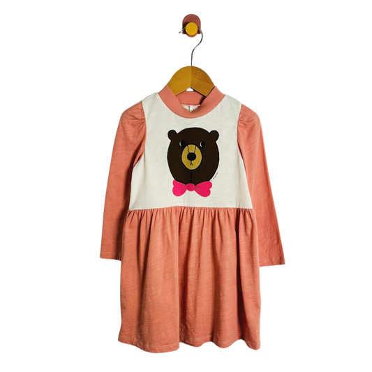 A toddler girl's long-sleeve dress with a terracotta orange skirt and sleeves, and a cream-colored bib-style chest panel featuring a cartoon bear face with a pink bow tie. The dress is hanging on a wooden hanger against a plain white background.