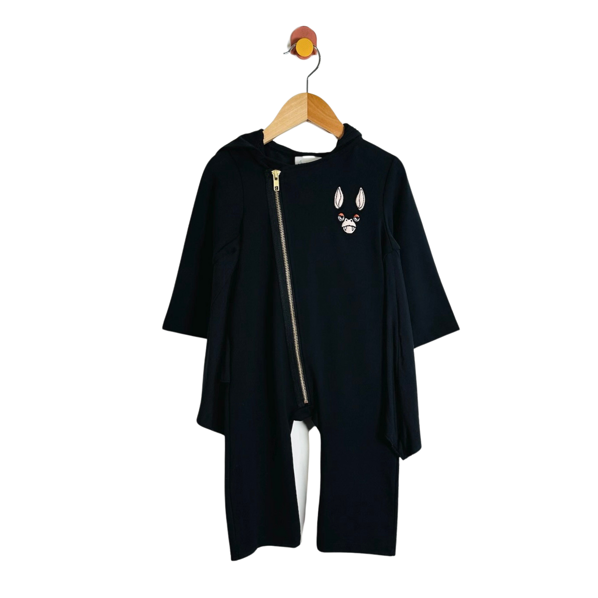 A black long-sleeve hooded oneise with an asymmetrical front zipper and a small embroidered bat face patch on the chest, hanging on a wooden hanger against a plain white background.
