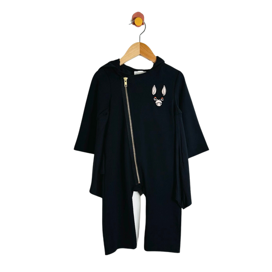 A black long-sleeve hooded oneise with an asymmetrical front zipper and a small embroidered bat face patch on the chest, hanging on a wooden hanger against a plain white background.