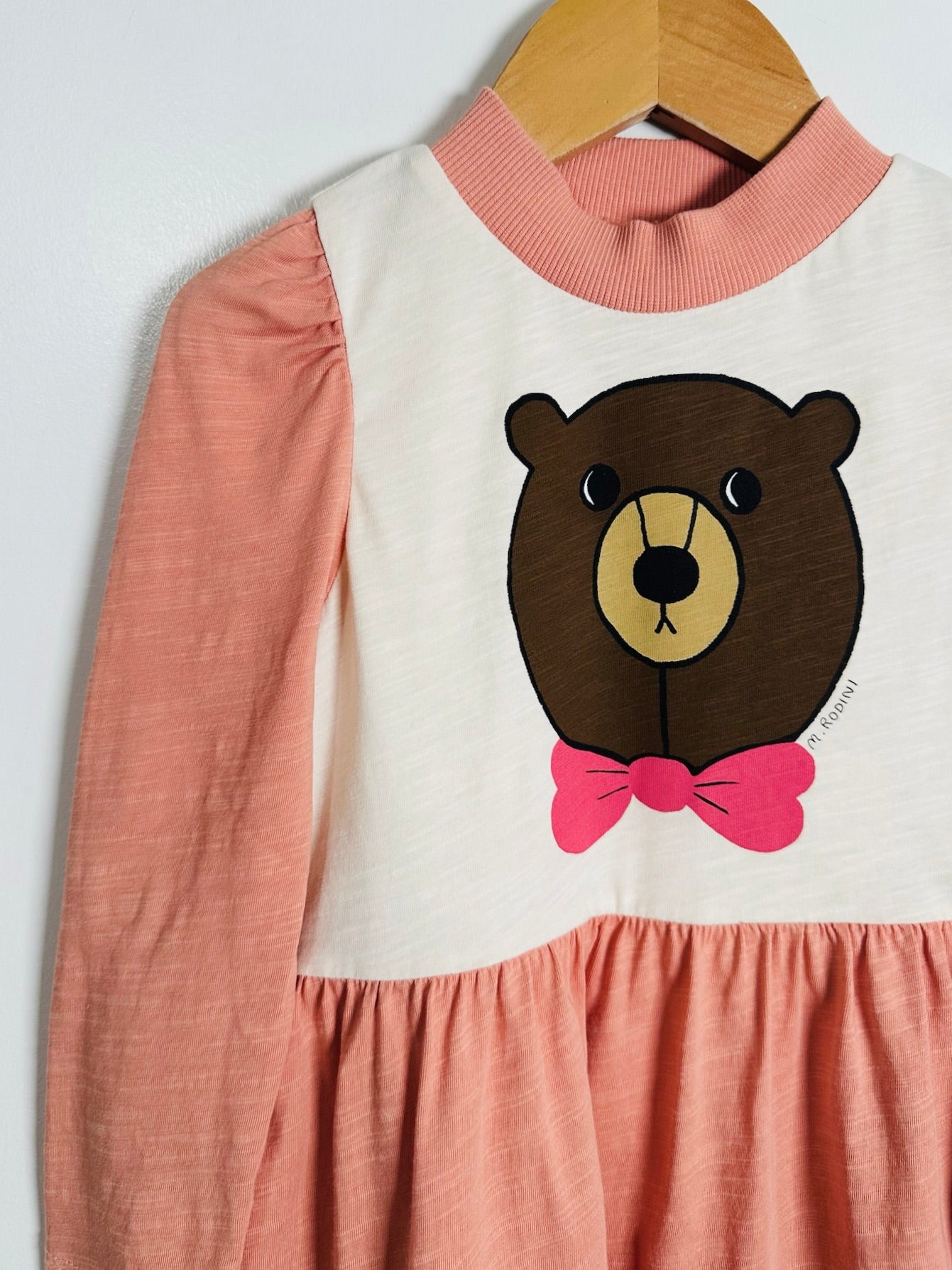 A children's long-sleeve dress hanging on a wooden hanger against a plain light background. The dress has a cream-colored upper bodice with a large brown bear face graphic and a bright pink bow tie, combined with a pink ribbed mock neck and a pink gathered skirt.