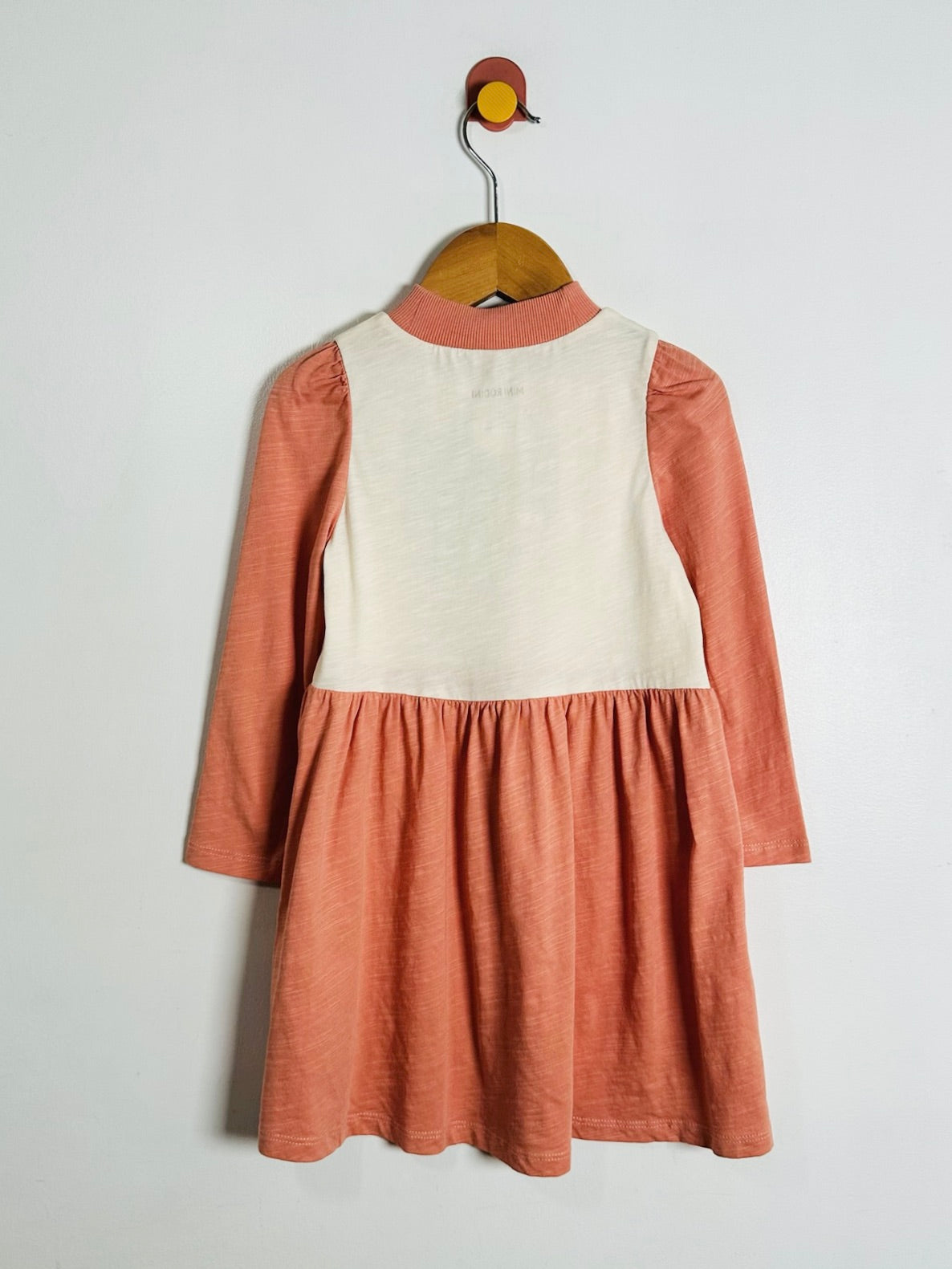 A children's dress hanging on a wooden hanger against a plain light gray wall. The dress has a warm peach-orange skirt and sleeves with a contrasting cream-colored front panel and a matching cream collar.