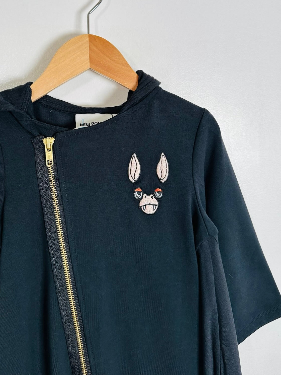 A dark navy blue zip-up hoodie with a gold zipper and an embroidered bat face on the chest, hanging on a wooden hanger against a plain white wall.