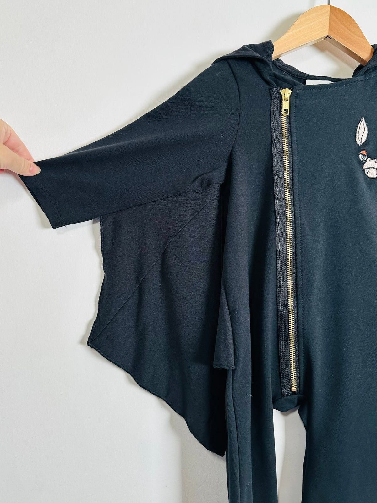 A black long-sleeve hooded oneise with an asymmetrical front zipper and a small embroidered bat face patch on the chest, hanging on a wooden hanger against a plain white background.