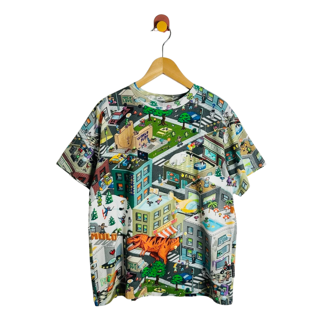 A short-sleeve crewneck t-shirt hanging on a wooden hanger against a plain white background. The t-shirt features a highly detailed, colorful isometric cartoon cityscape print with various buildings, roads, and characters.