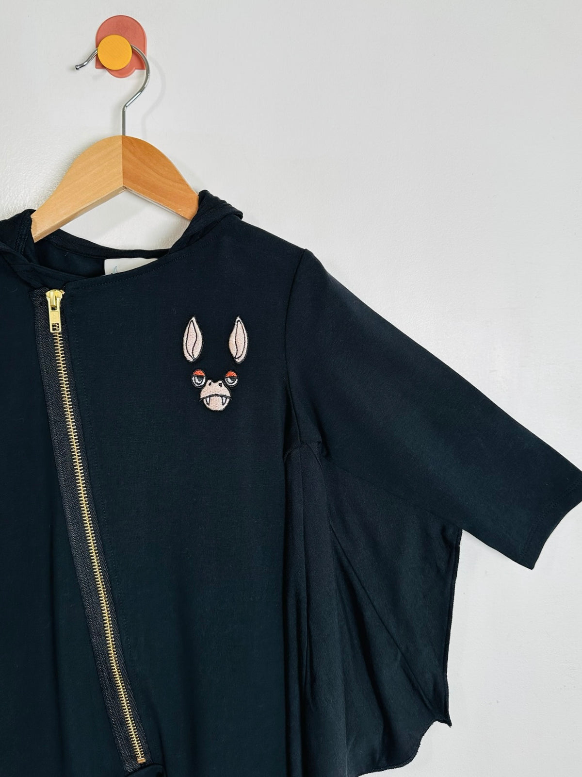 A black short-sleeve zip-up hoodie with a gold zipper and a white bat face graphic on the chest, hanging on a wooden hanger against a light gray wall.
