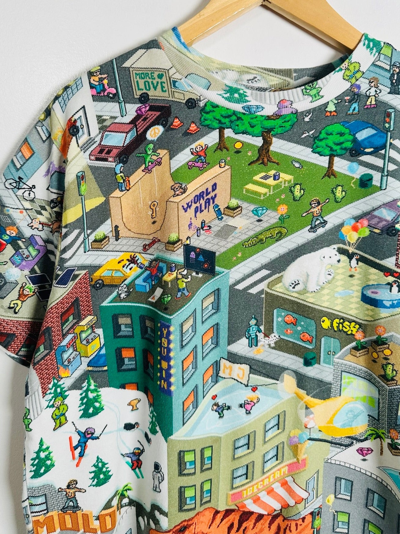 A short-sleeve crewneck t-shirt hanging on a wooden hanger, featuring a highly detailed, colorful cartoon-style city street scene with buildings, people, trees, and vehicles.