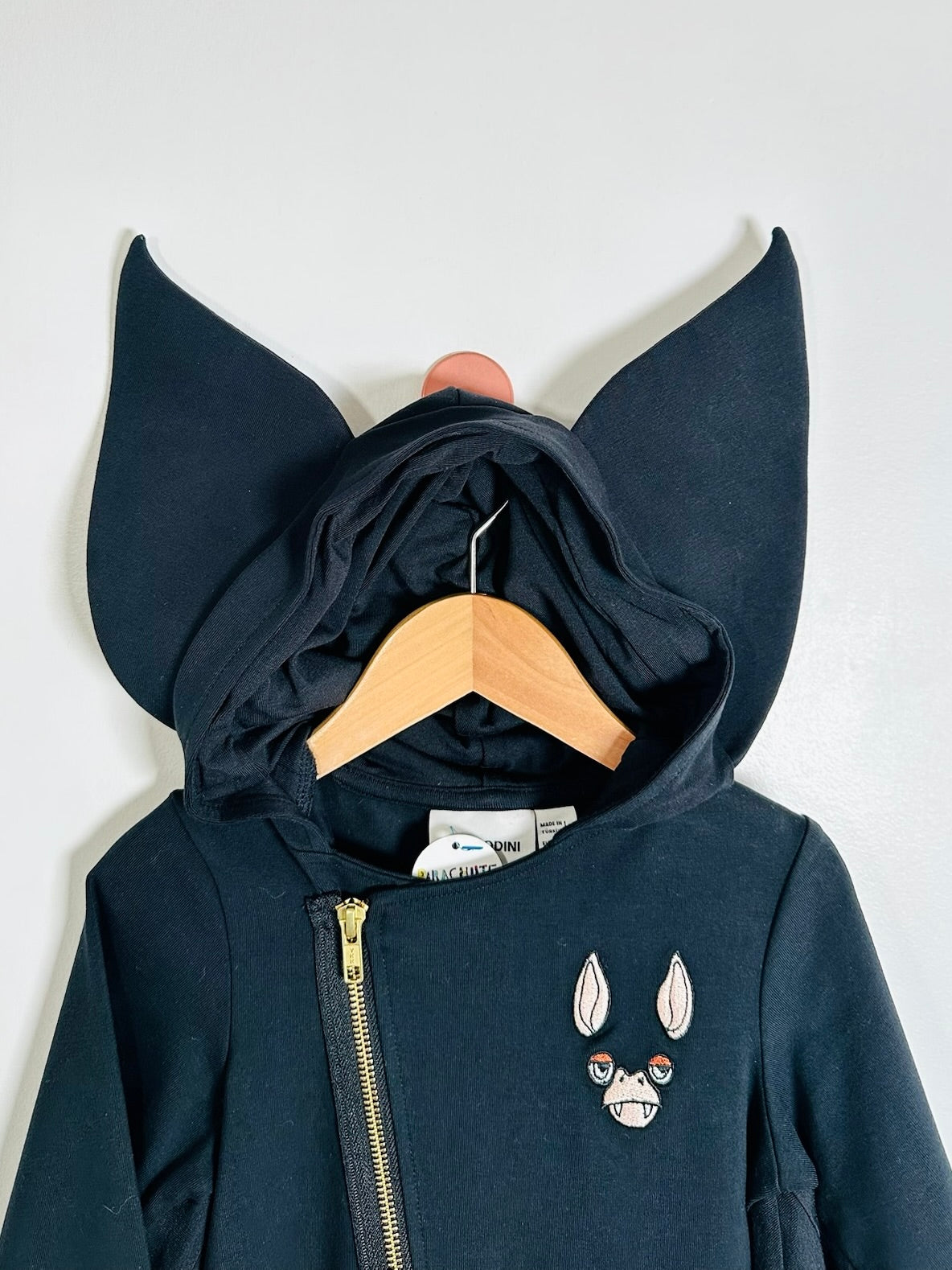 A dark navy blue zip-up hoodie with large pointed bat-ear extensions from the hood, hanging on a wooden hanger against a plain light gray wall. The hoodie features a gold zipper, a small embroidered cat face patch on the left chest, and a white brand tag inside the collar.