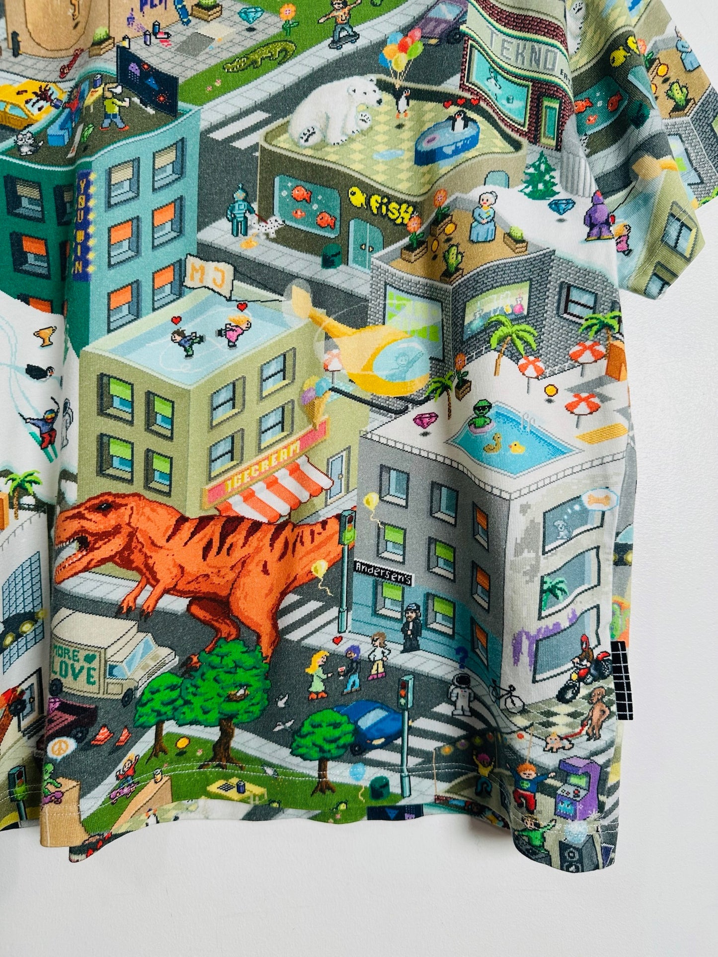 A close-up of a multicolor printed fabric featuring a whimsical illustrated city scene with buildings, people, cars, trees, and a large orange dinosaur, shown draped against a light gray background.