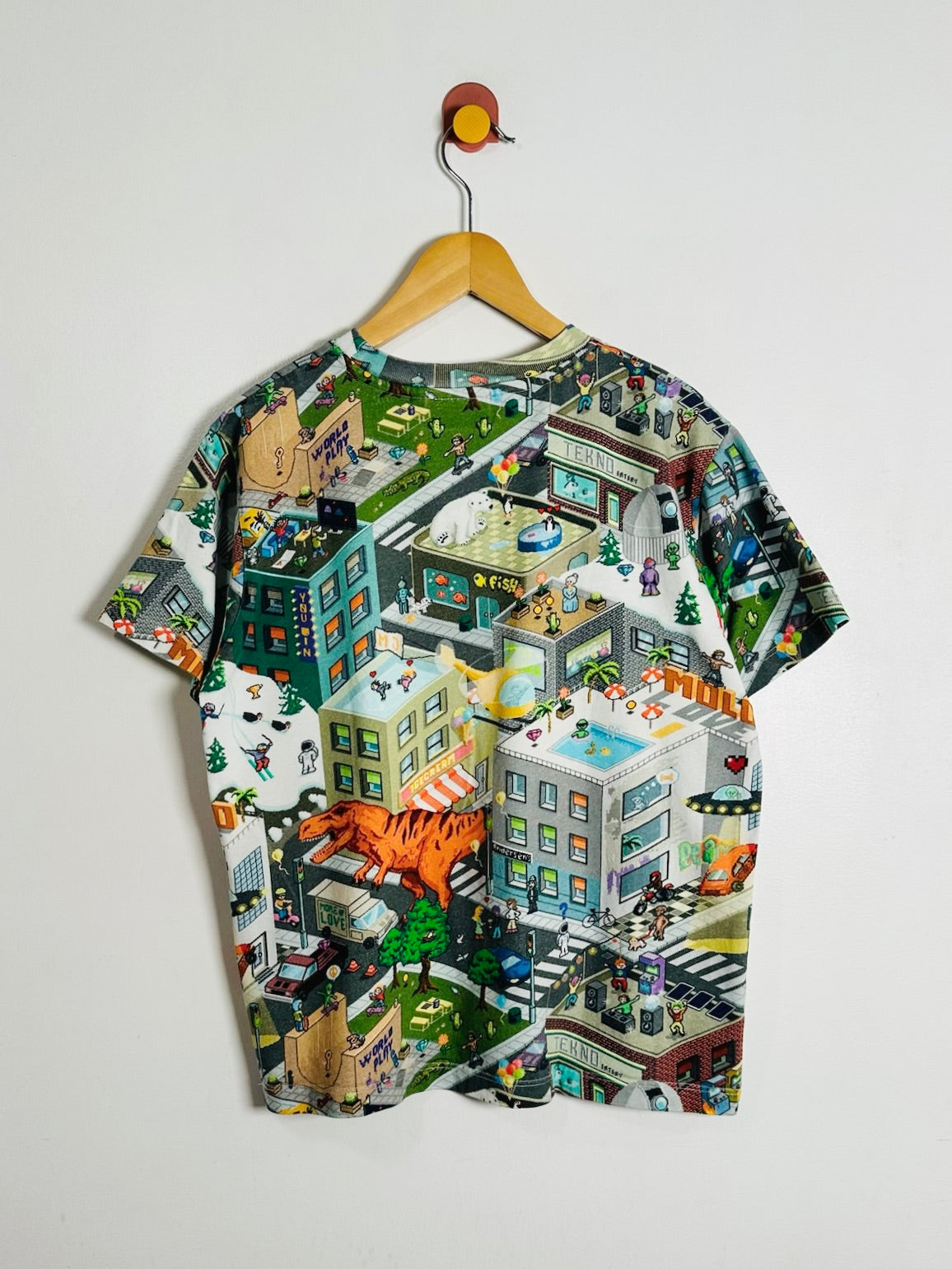 A short-sleeve crewneck t-shirt with a highly detailed, colorful isometric cityscape graphic print, hanging on a wooden hanger against a plain white wall.
