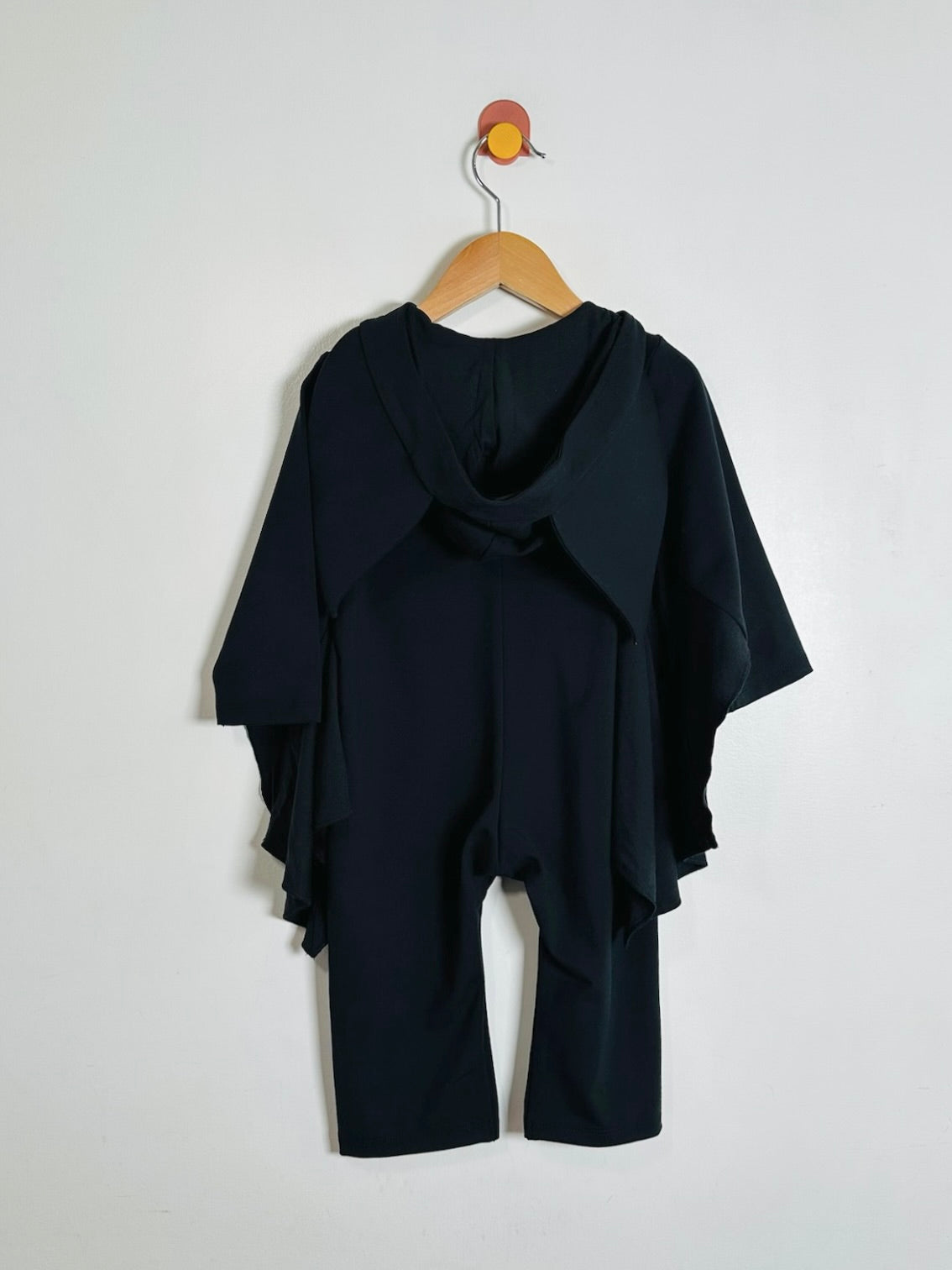 A black long-sleeve hooded oneise with an asymmetrical front zipper and a small embroidered bat face patch on the chest, hanging on a wooden hanger against a plain white background.