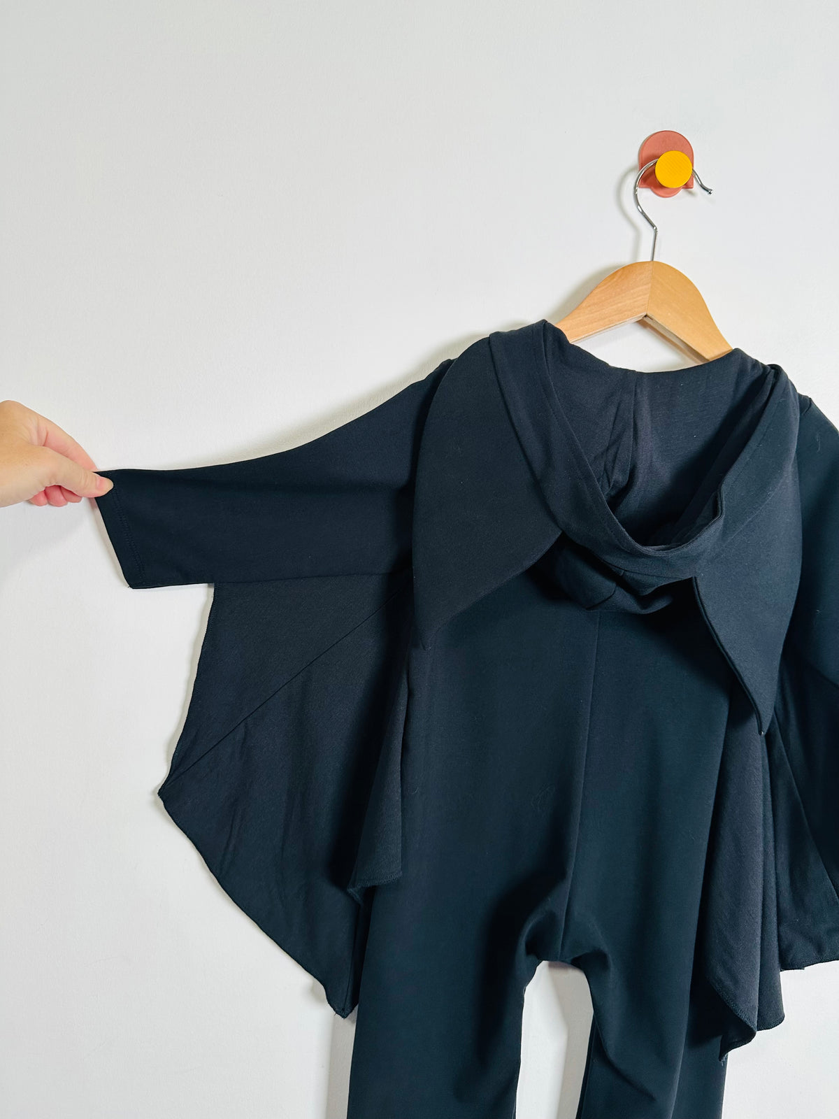 A black long-sleeve hooded oneise with an asymmetrical front zipper and a small embroidered bat face patch on the chest, hanging on a wooden hanger against a plain white background.