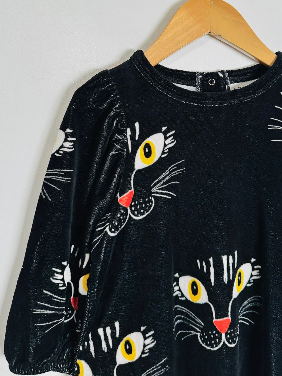 A black long-sleeve top with a playful cat-face print, hanging on a wooden hanger against a plain light gray wall.