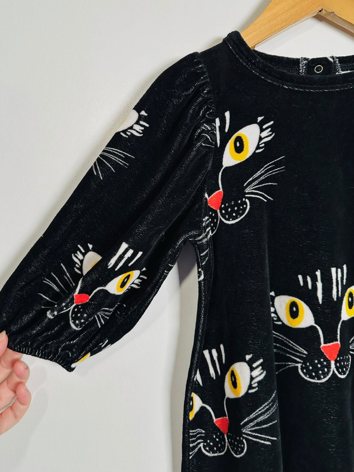 A black velvet long-sleeve top with a playful cat-face print, hanging on a wooden hanger against a plain white wall. A hand is visible holding the sleeve.