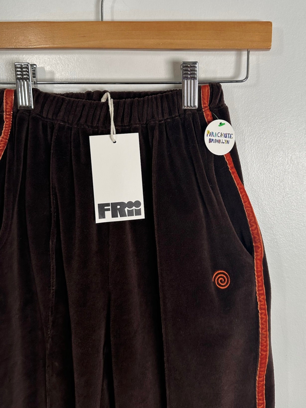 Dark brown velour pants with orange side stripes and a small orange spiral logo, hanging on a wooden hanger with a white brand tag and a circular size tag.