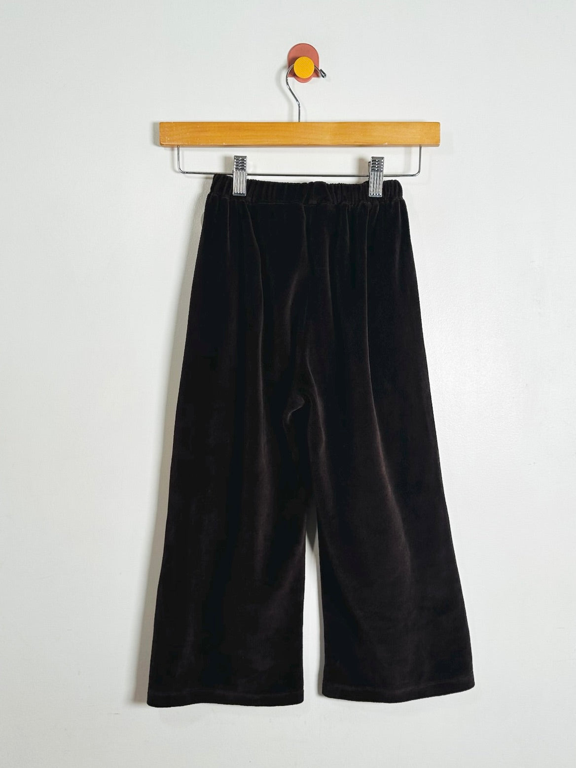 Black wide-leg velvet pants hanging from a wooden hanger against a plain white wall.