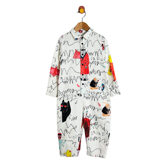 A children's long-sleeve one-piece jumpsuit with a white base and an all-over graffiti-style black scribble print, featuring colorful cartoon character illustrations in red, yellow, blue, and black. The jumpsuit is shown hanging from a wooden hanger with a bird hook against a plain white background.