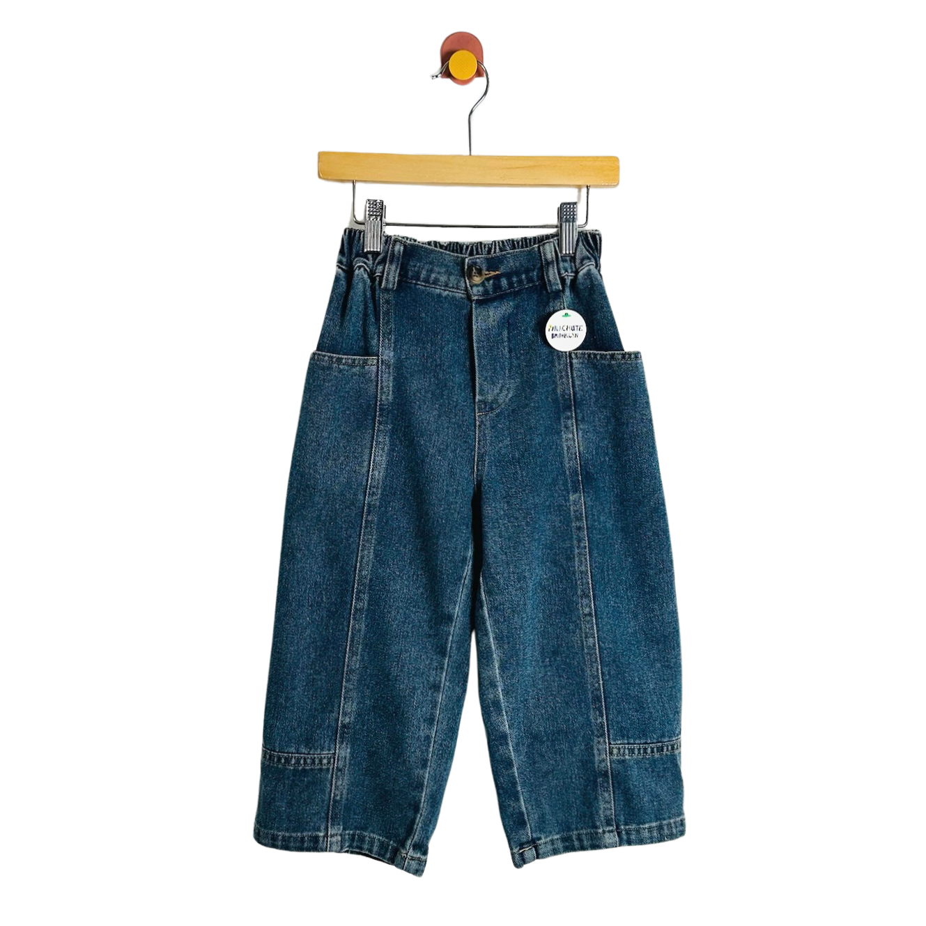A pair of wide-leg blue denim pants with front patch pockets, hanging from a wooden hanger against a plain white background.
