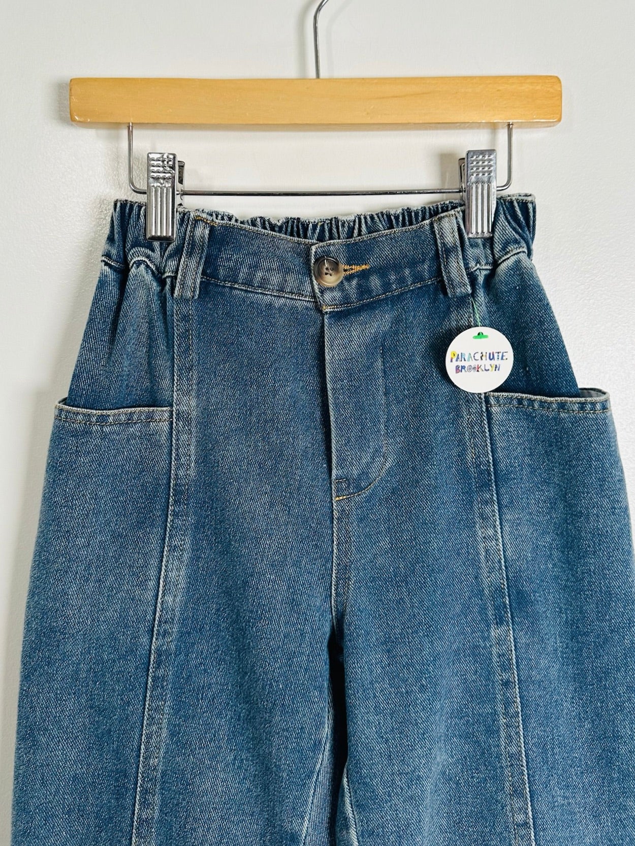 A pair of blue denim jeans with a high waist and front patch pockets, hanging from a wooden hanger against a white wall. A round white tag is attached to the right side of the waistband.