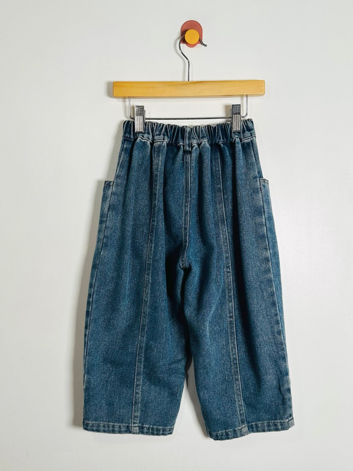 A pair of blue denim pants with an elastic waistband and side pockets, hanging from a wooden hanger against a plain light gray wall.
