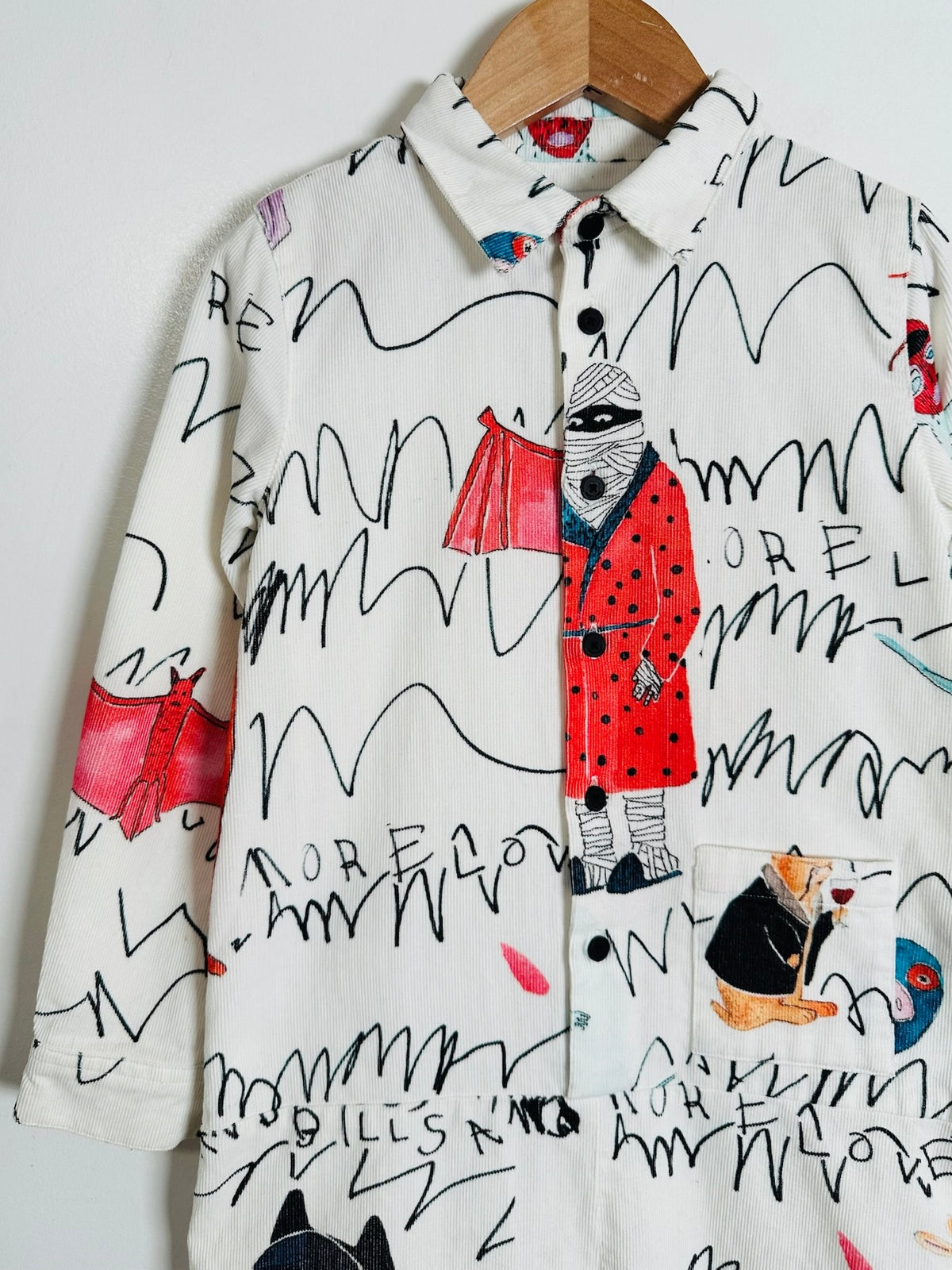 A long-sleeve button-up shirt on a wooden hanger, featuring an off-white base with black scribble lines and whimsical illustrations of figures in red and black, including one holding a red bag.