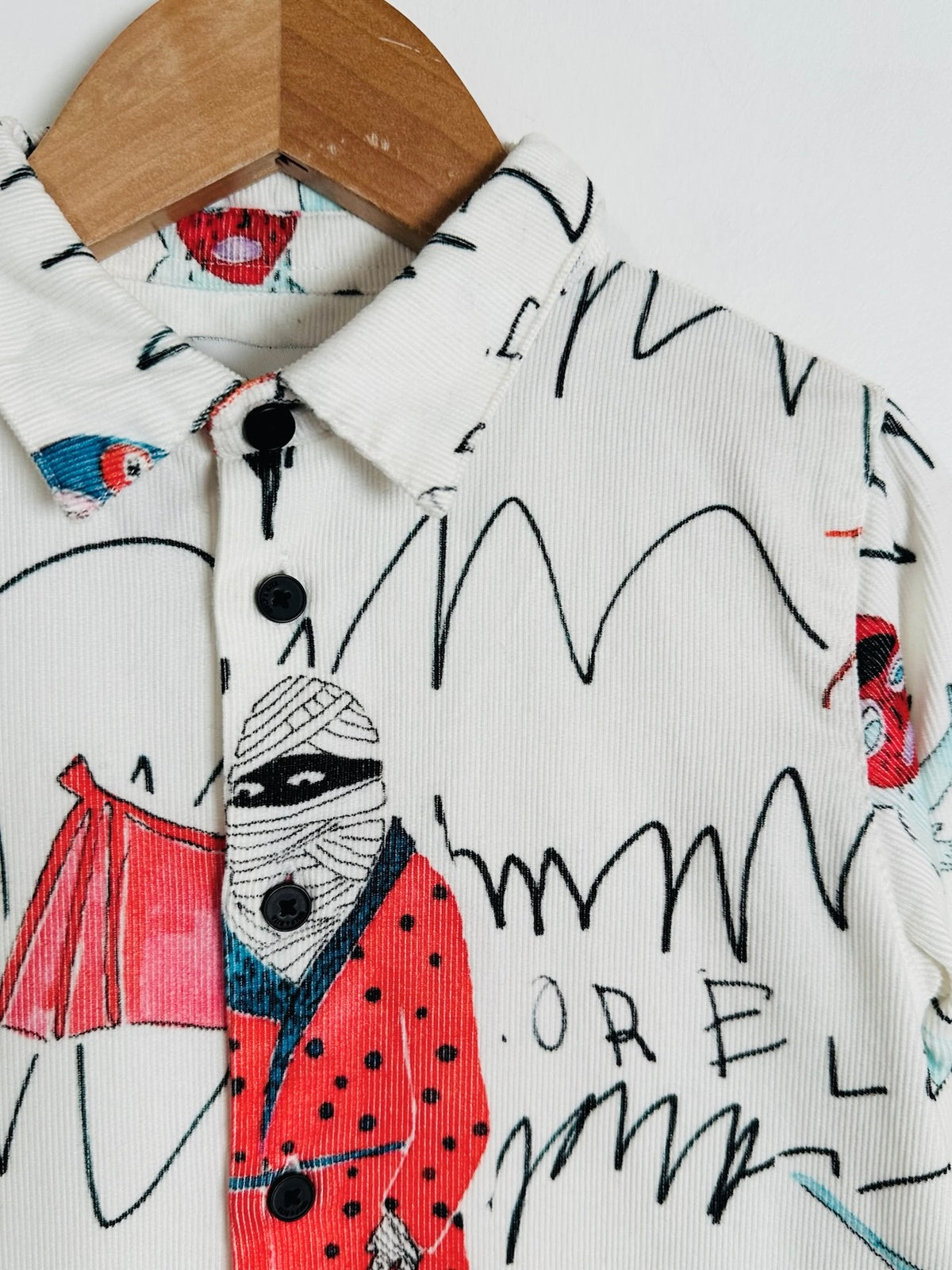 A close-up of a white corduroy button-up shirt on a wooden hanger, featuring a whimsical graffiti-style illustration of a bandaged figure in a red polka-dot coat and black scribbled lines.
