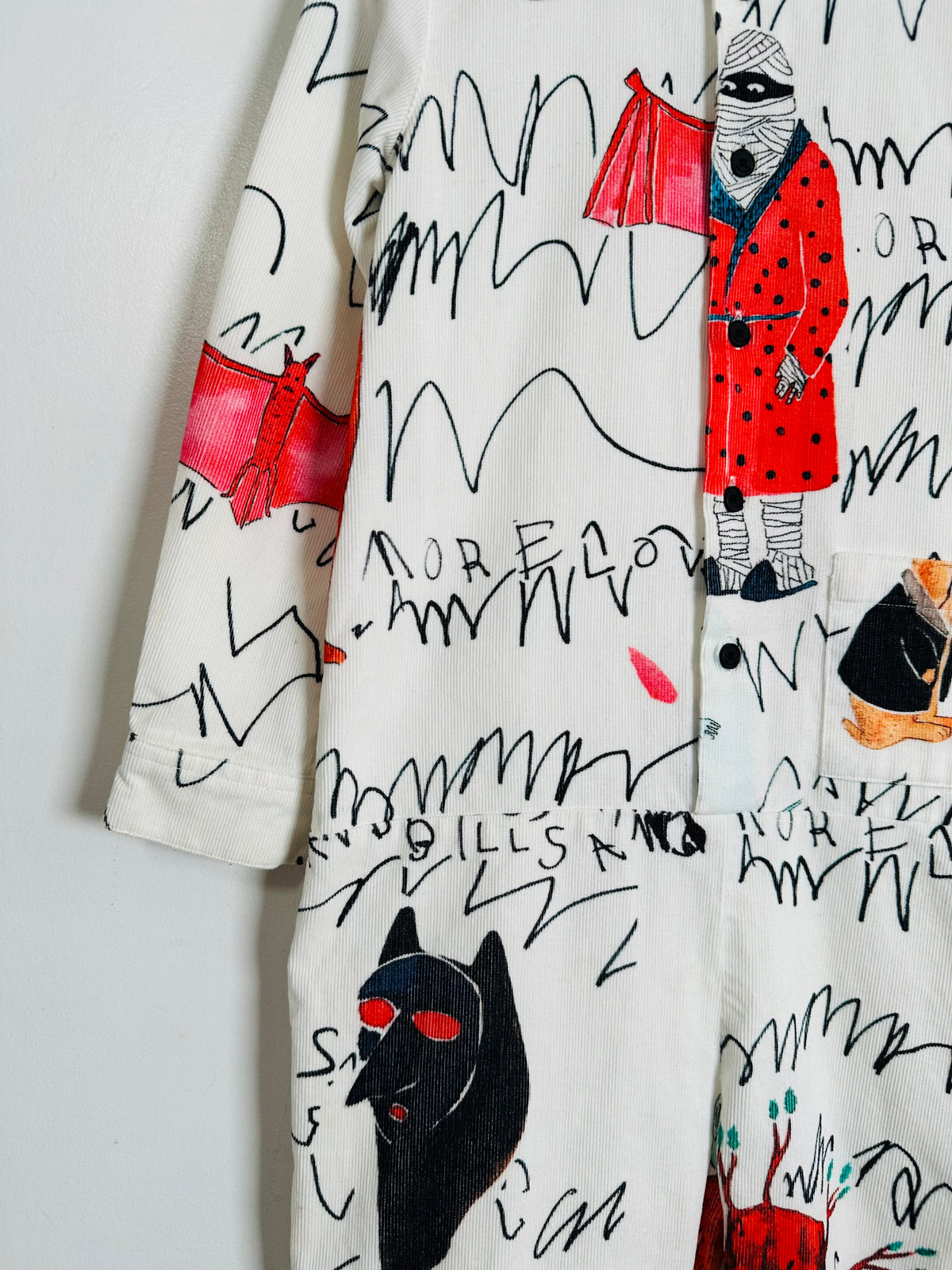 A close-up of a white long-sleeve garment featuring a whimsical, hand-drawn style print with black scribbles, a red hooded figure, a black cat with red eyes, and text elements.