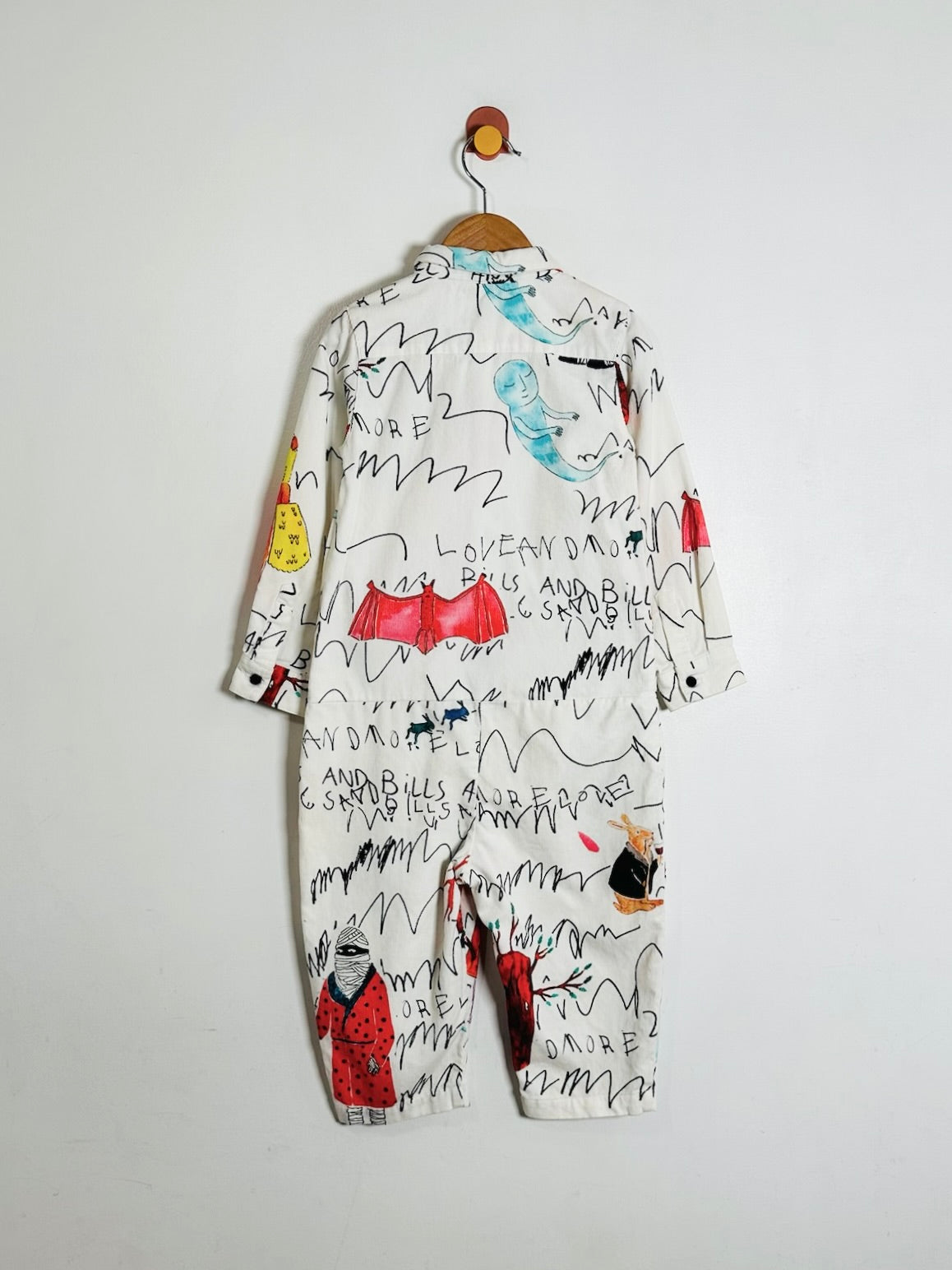 A long-sleeve baby romper in white, decorated with a playful black scribble pattern and colorful illustrations of cartoon figures and text, hanging on a wooden hanger against a plain white wall.