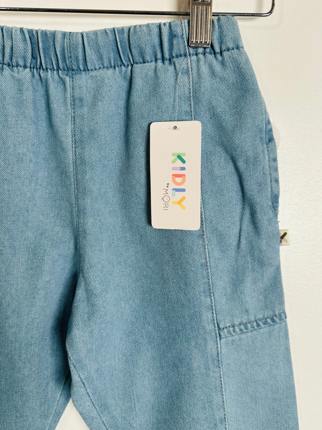 Light blue denim pants hanging on a metal hanger against a white wall, with a white hangtag that reads KidlY visible.