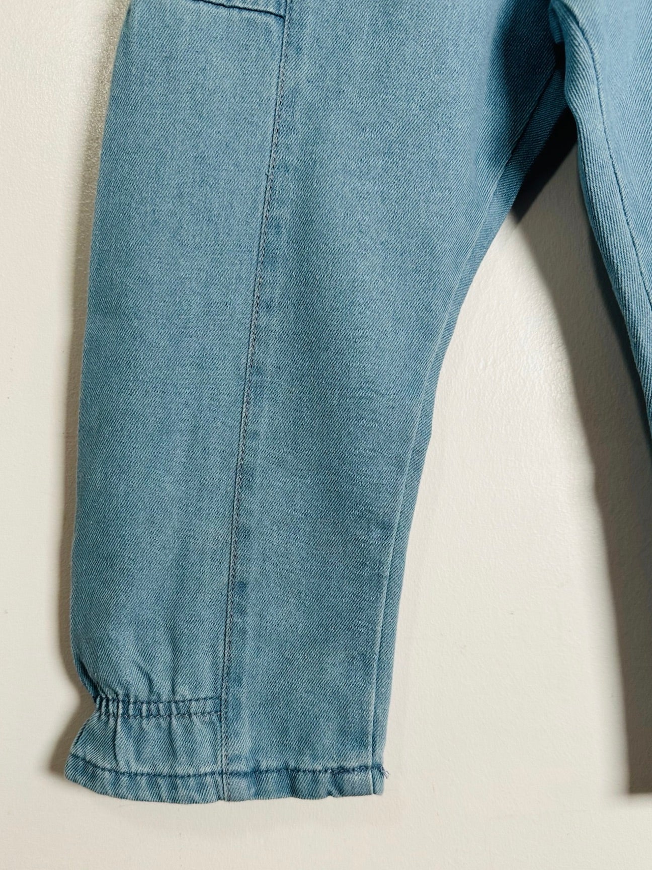 Close-up of a light blue denim garment, showing the fabric texture, seam, and elasticized cuff detail against a plain off-white background.