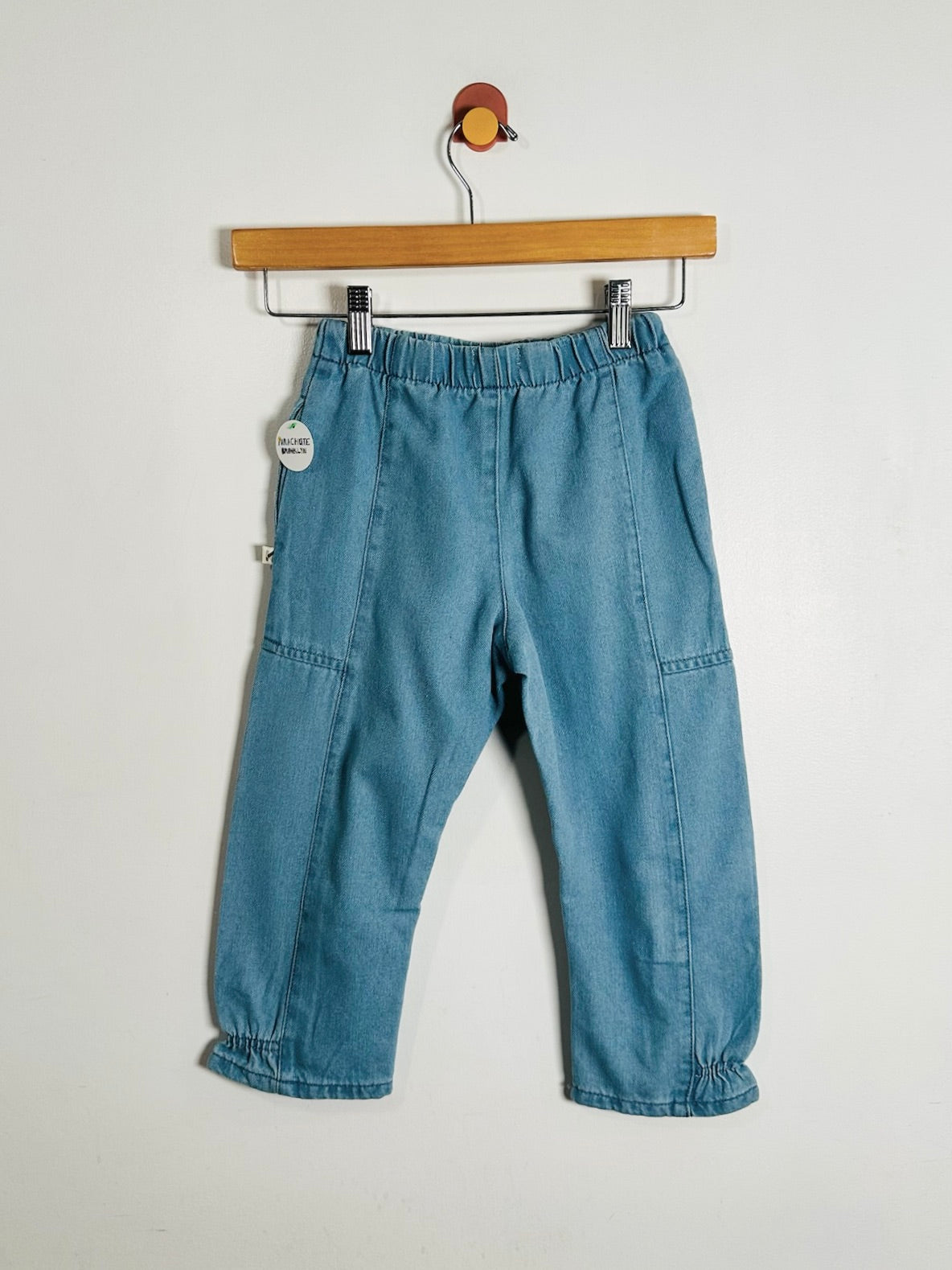 Light blue denim pants for toddlers, hanging from a wooden hanger against a plain off-white wall. The pants feature an elastic waistband, side pockets, and gathered cuffs.