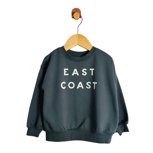 A dark slate green crewneck sweatshirt with 'EAST COAST' printed in white block letters on the chest, hanging on a wooden hanger against a plain white background.
